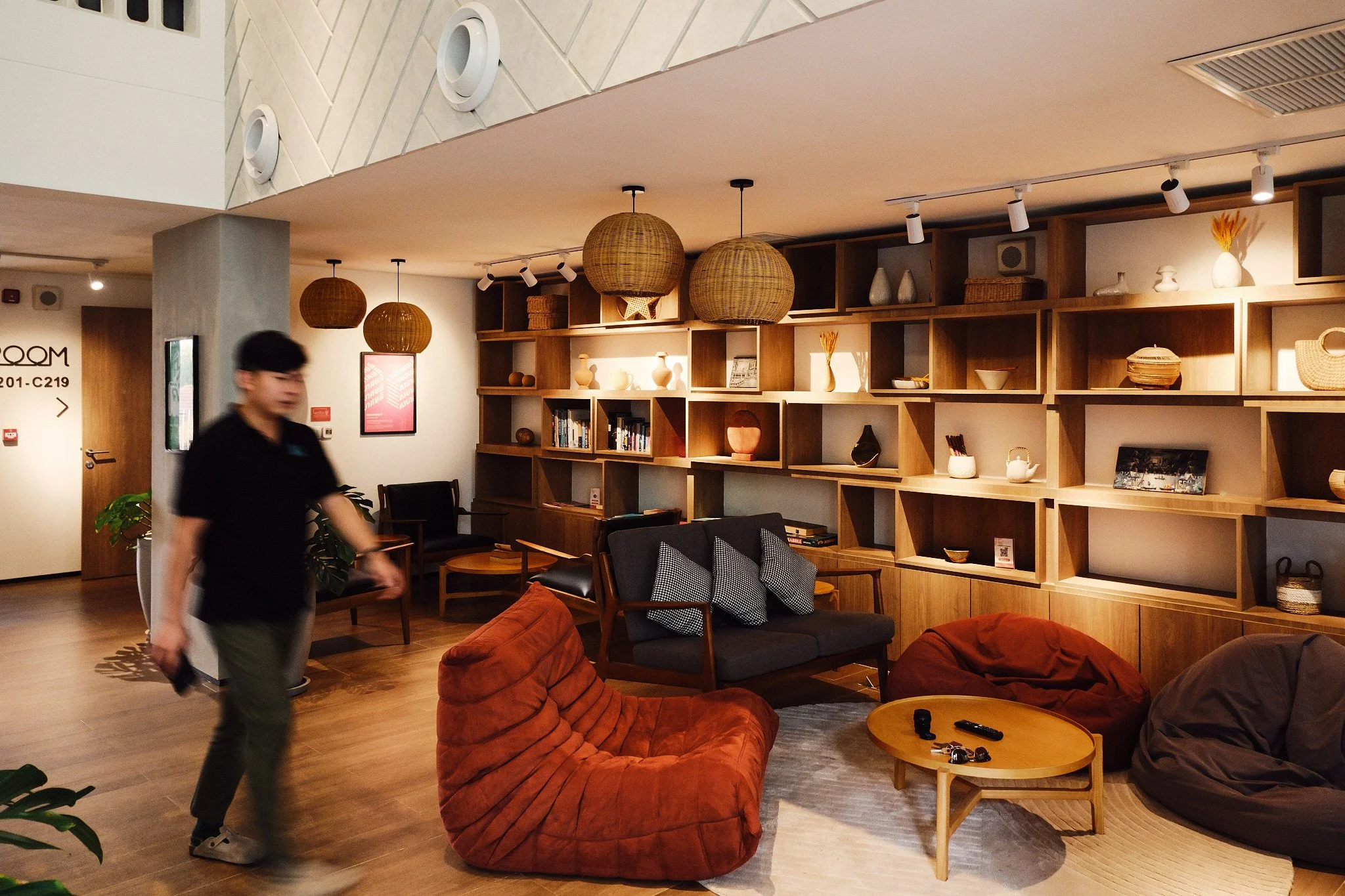 Interior of a modern lounge or waiting area with wooden shelving filled with decorative items and books, wicker pendant lights, comfortable chairs and sofas, and a person walking through the space.