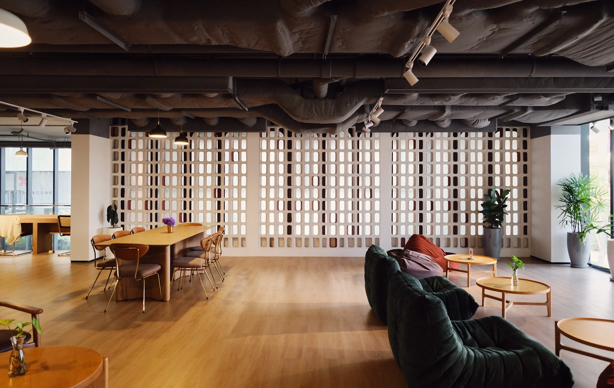 A spacious modern indoor lounge area with large windows, potted plants, and comfortable seating including armchairs and couches, with a decorative partition in the background and ambient lighting.