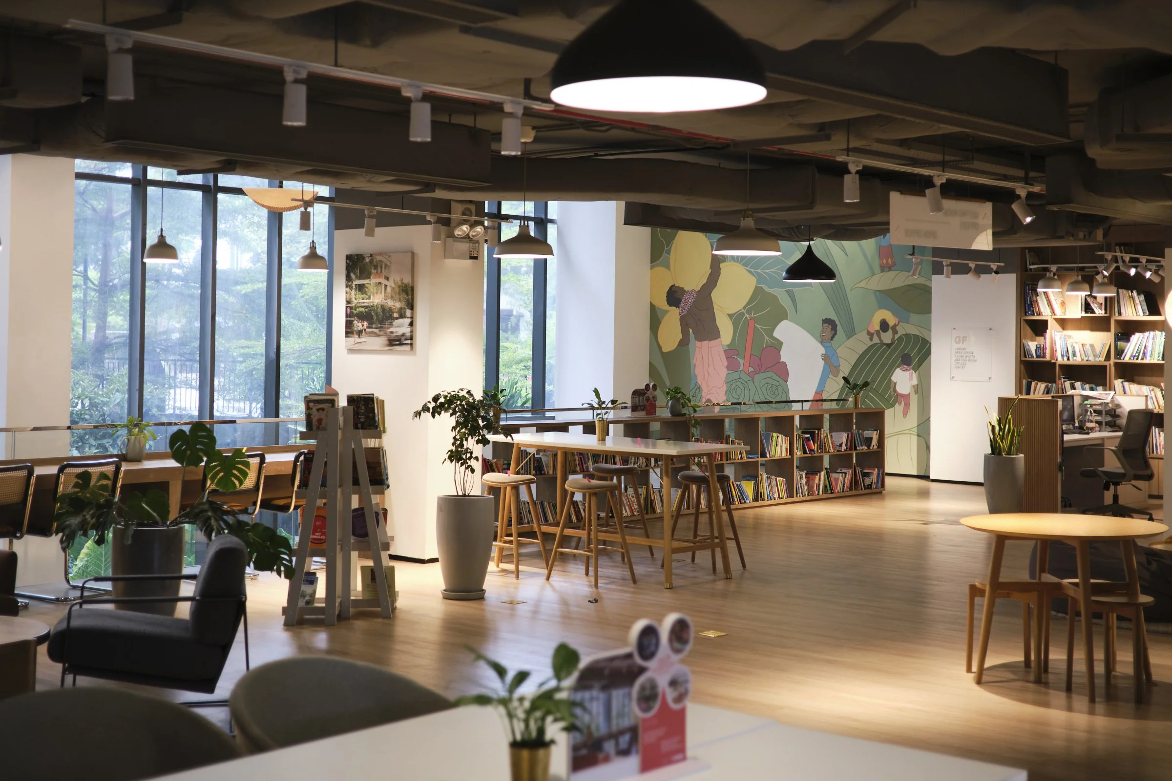 Interior of a modern library or bookstore with large windows, wooden floors, multiple bookshelves, and seating areas. Plants and decorative artwork on the walls are visible, along with hanging lights and a mural of people and large flowers.