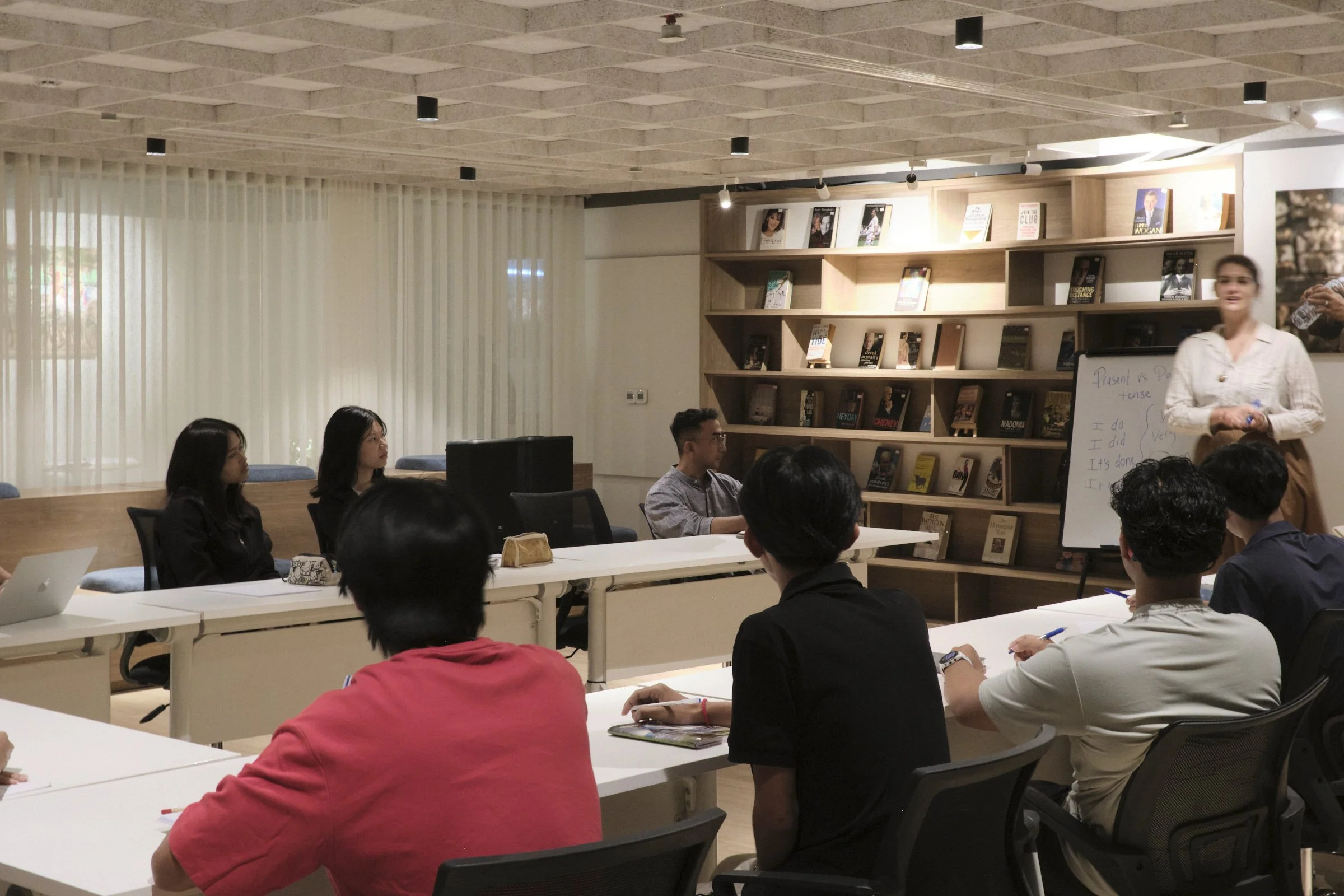 A group of people attending a presentation or workshop in a modern conference room with bookshelves on the wall. A woman in a light-colored blouse is standing near a whiteboard, speaking to the audience.