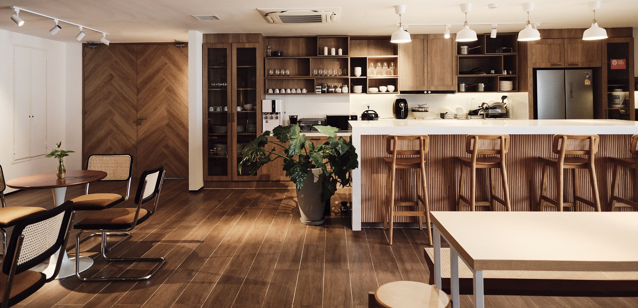 Modern kitchen and dining area with wooden furniture, bar stools at a white counter, open shelving with dishes, and a potted plant.