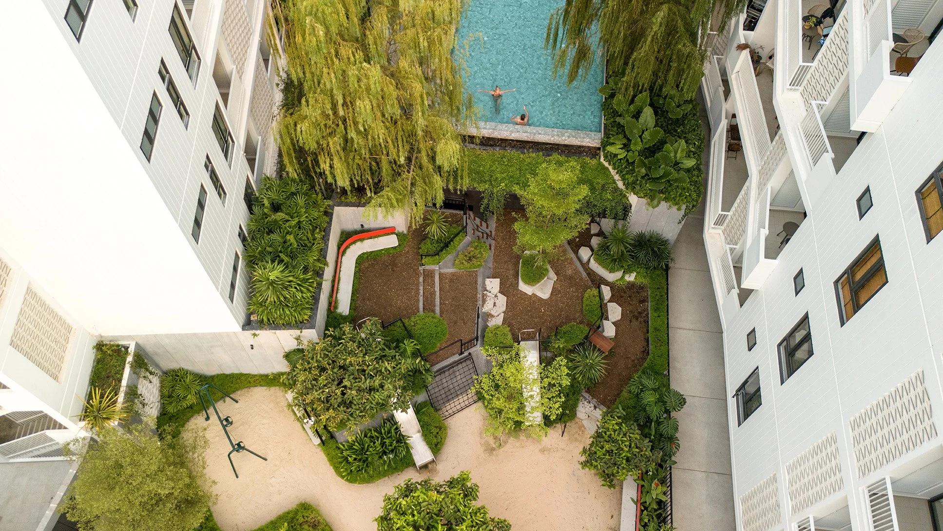 Aerial view of a residential apartment courtyard featuring a swimming pool with two people swimming, surrounded by lush green trees, landscaped bushes, paved walkways, a small garden area, and white apartment buildings with balconies.