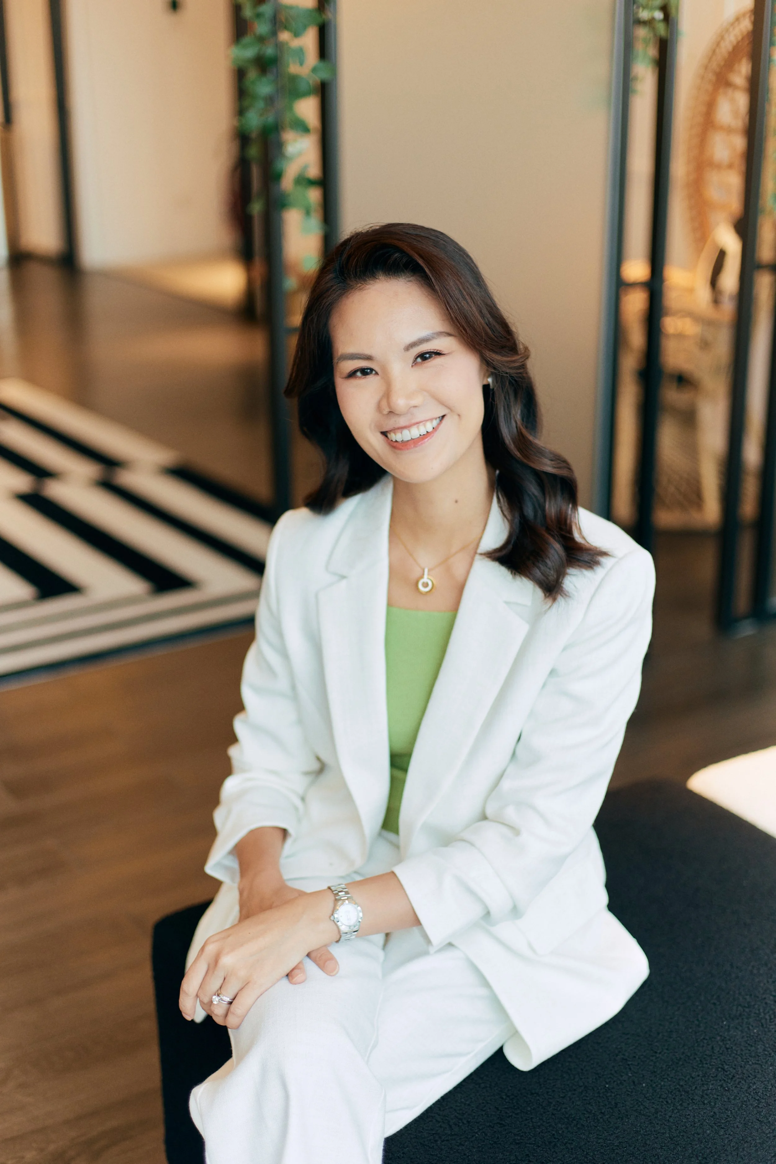 A smiling woman with dark wavy hair, wearing a white blazer and pants, light green top, and jewelry, sitting on a black cushion in a modern, stylish indoor setting.