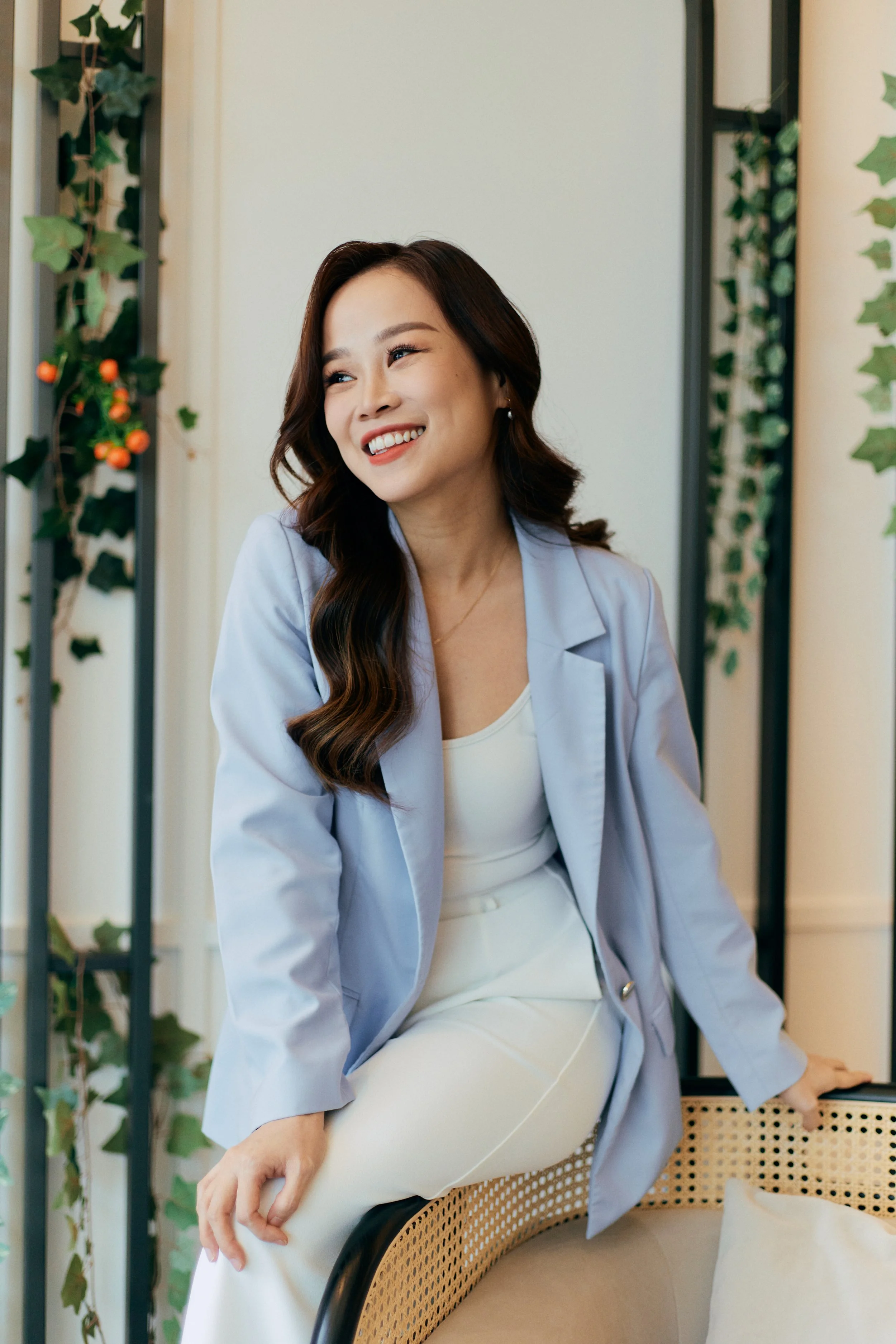 A woman with long wavy dark hair and light skin, dressed in a white top and cream-colored pants, is sitting on a rattan bench with a black frame. She is wearing a light blue blazer and smiling, looking to her right.