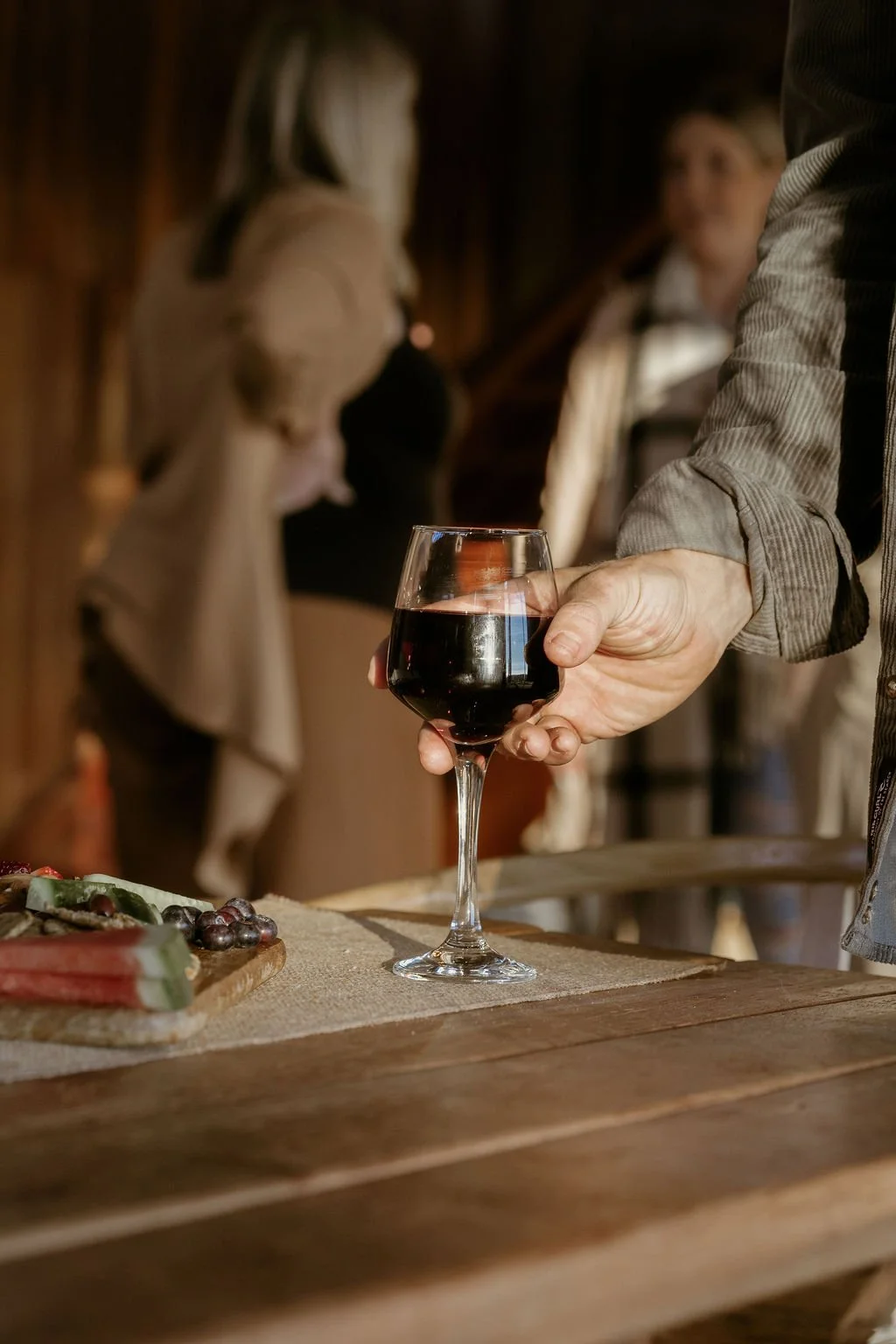 Person holding a glass of red wine over a wooden table with a charcuterie board, in a cozy indoor setting with other people in the background.