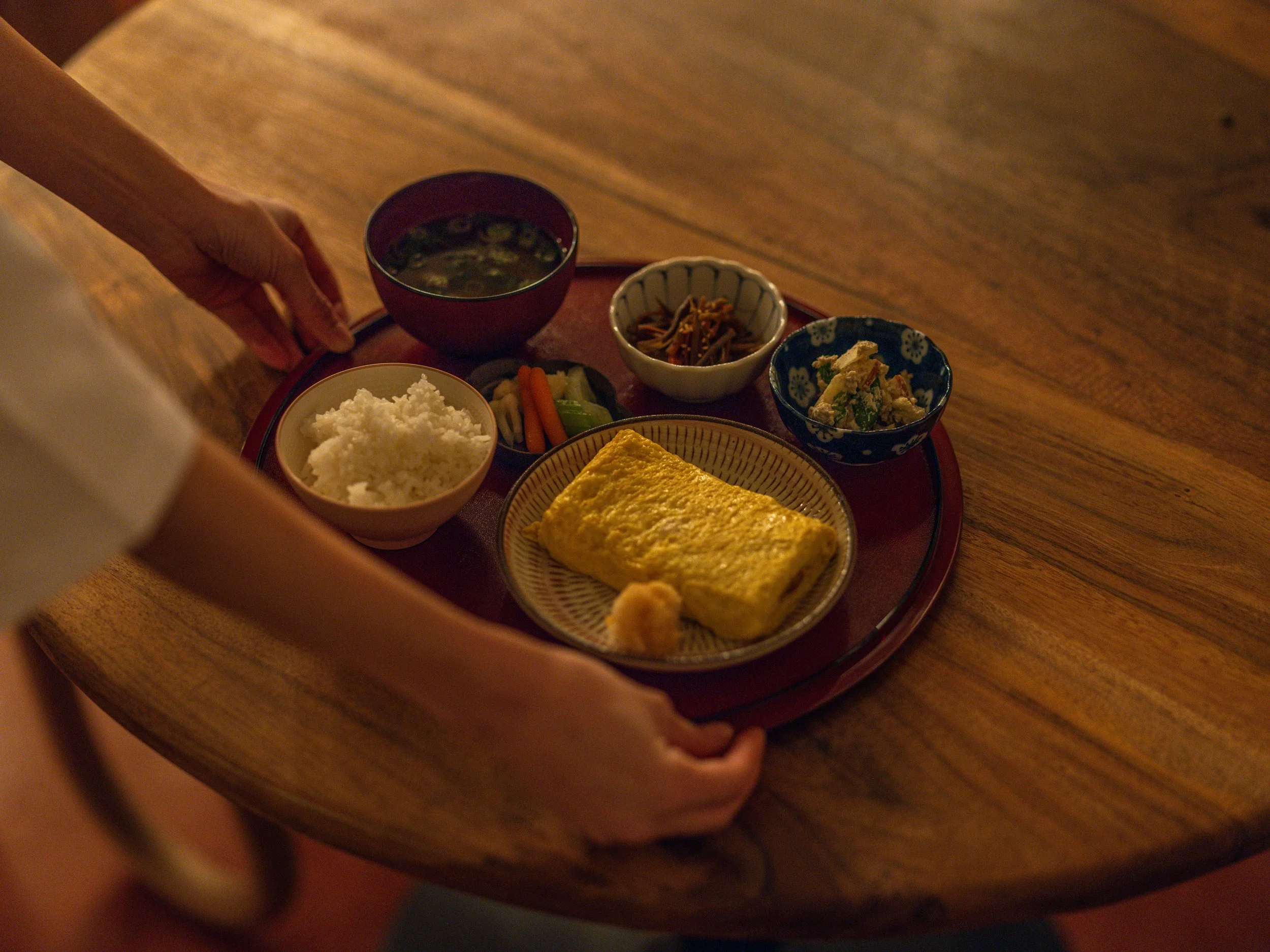A tray of traditional Japanese breakfast with rice, omelette, miso soup, and various side dishes on a wooden table.