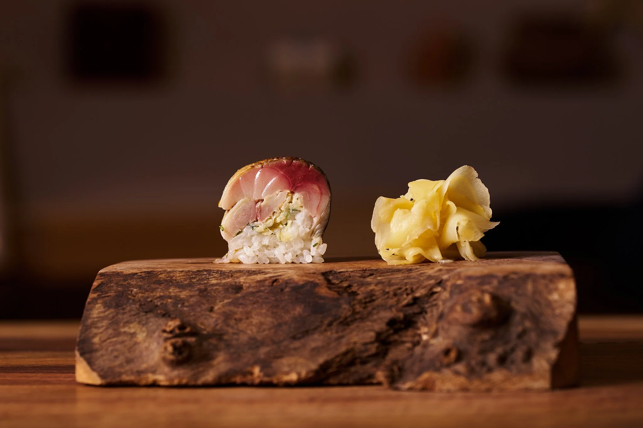 A piece of sushi with fish and rice on a wooden block, accompanied by a pile of pickled ginger.