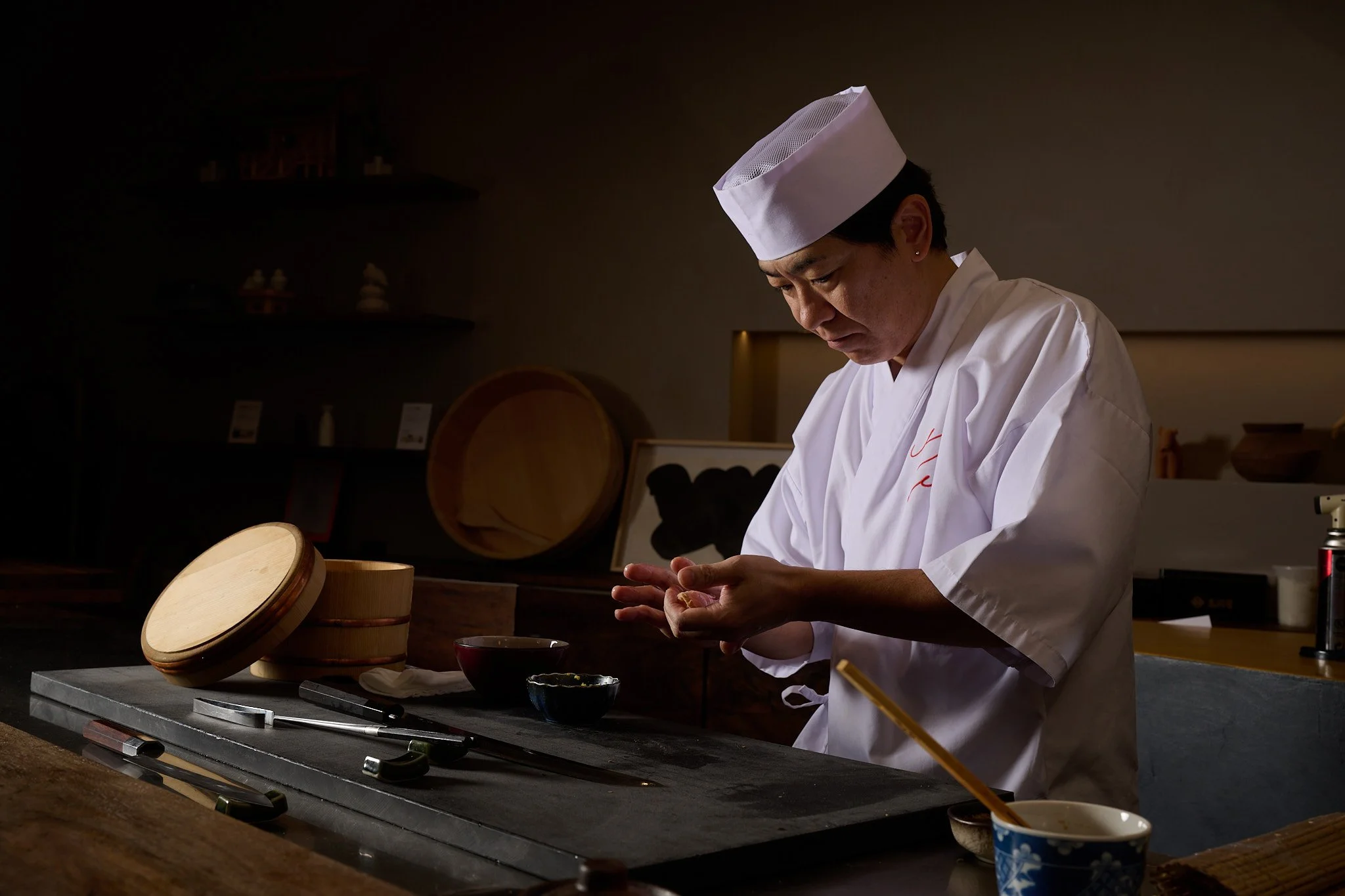 A chef in traditional white attire and hat preparing food in a dimly lit kitchen.