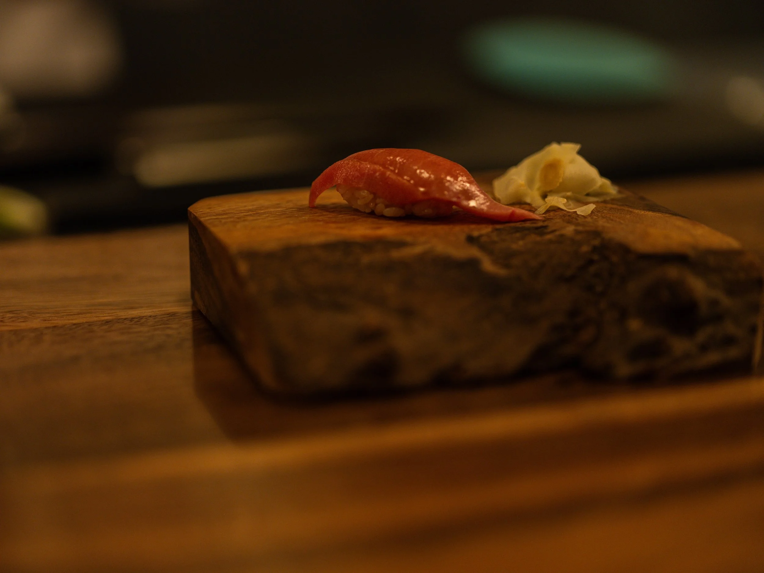 Close-up of a piece of sushi with pink fish and rice on a wooden plate, with a small amount of wasabi or horseradish beside it, in a dimly-lit setting.
