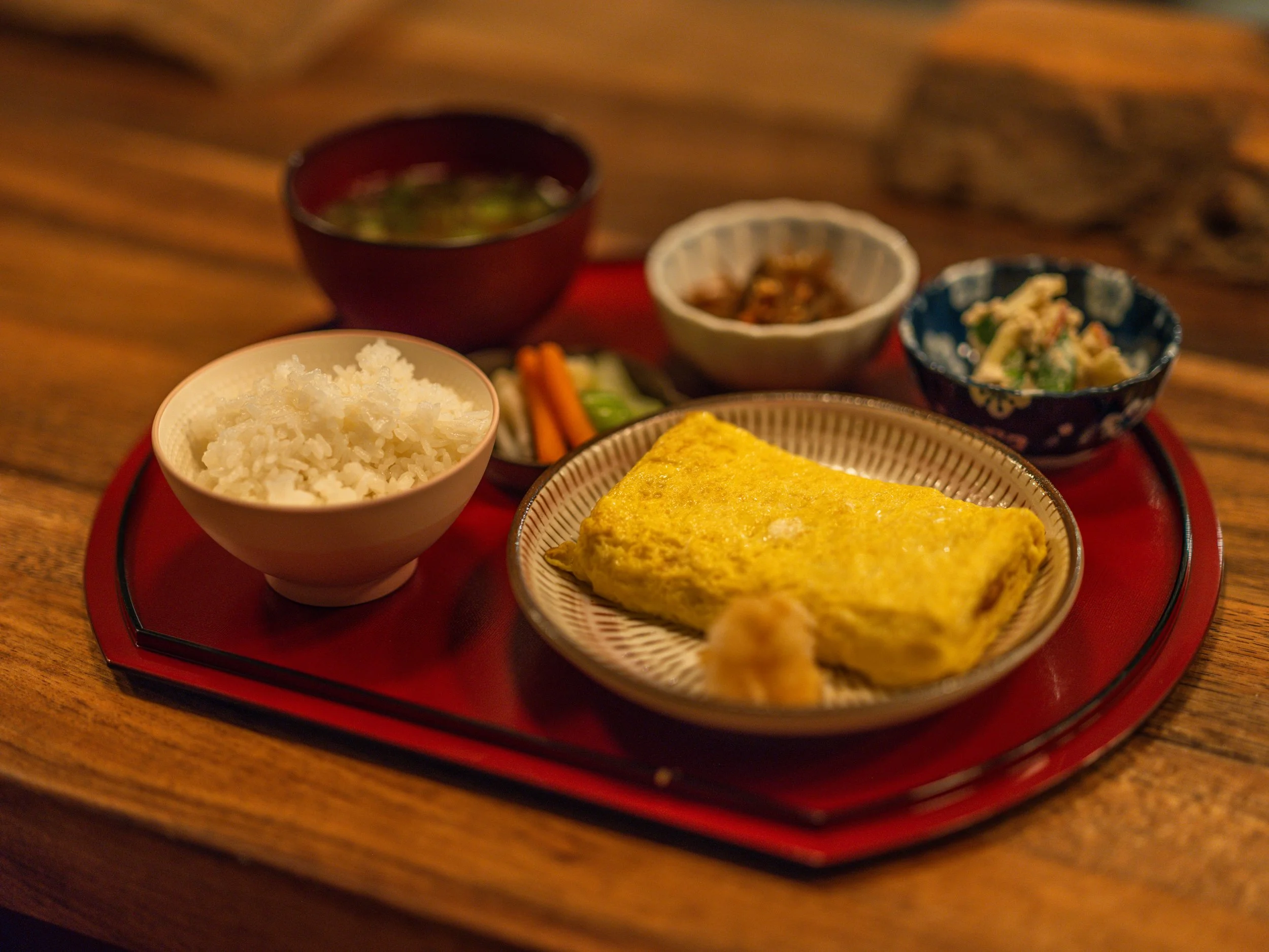 A traditional Japanese breakfast plate with rice, an omelette, pickled vegetables, and small side dishes including soup and salad on a red tray.