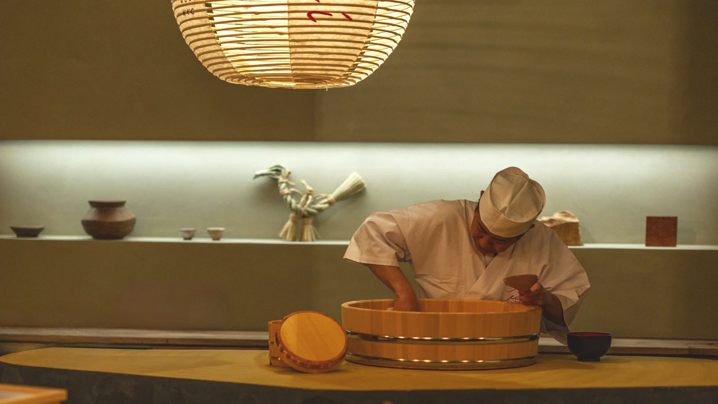 A chef in traditional white attire and hat preparing food in a Japanese-style kitchen. The scene is illuminated by a large paper lantern and features wooden bowls and utensils.