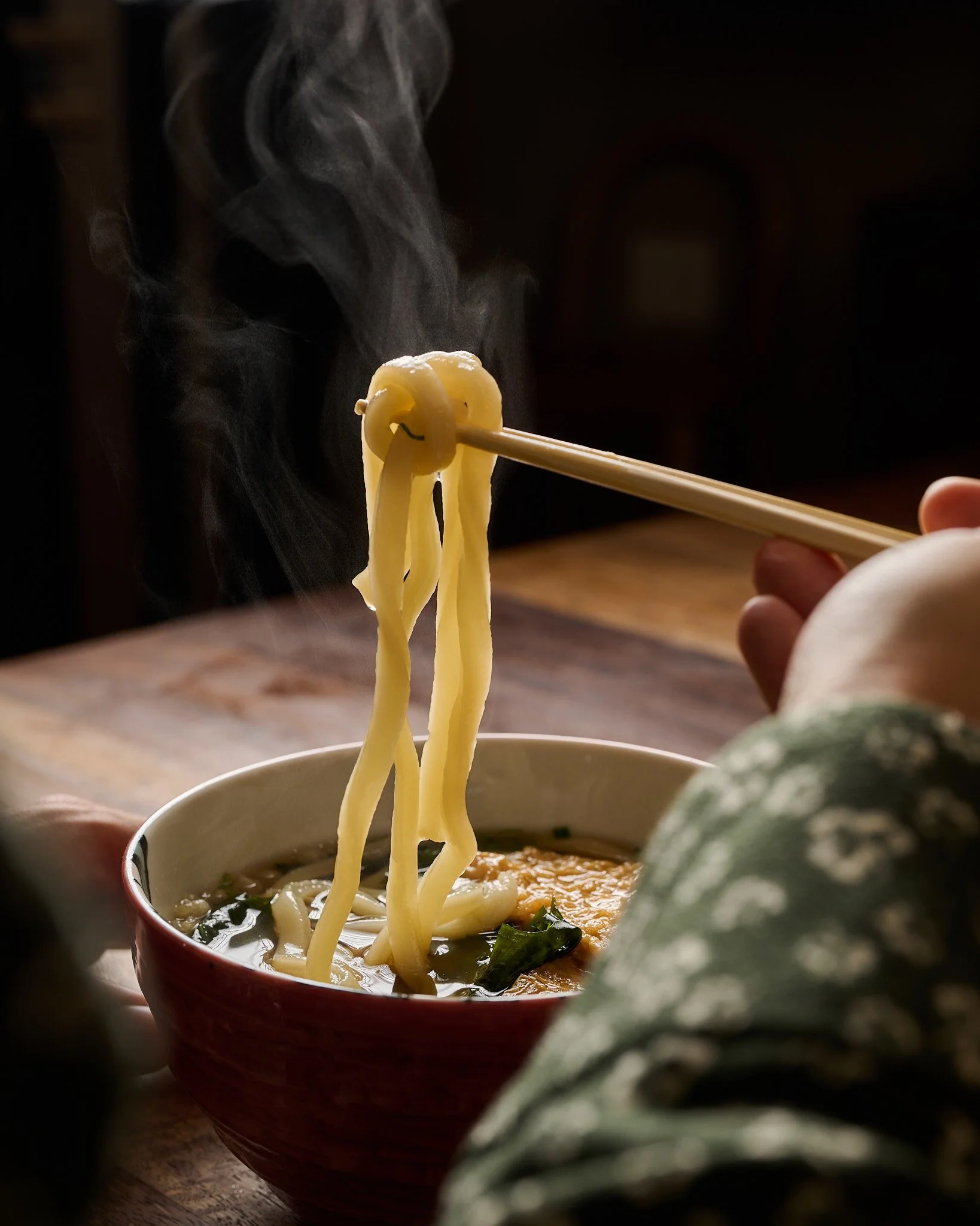 Person holding chopsticks lifting noodles from a steaming bowl of noodle soup, with steam rising, on a wooden table.
