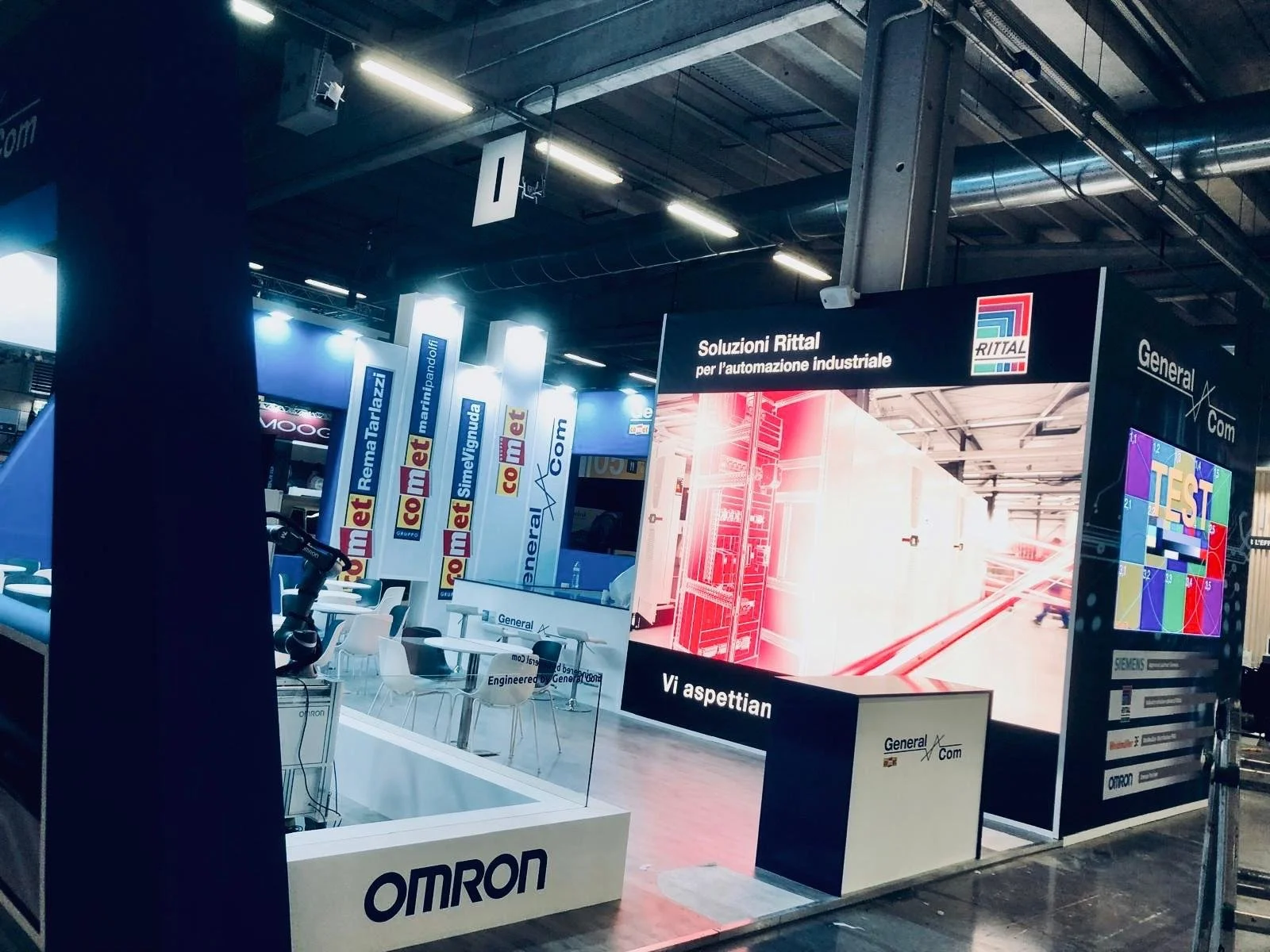 Stand espositivo di Omron con schermo e pannelli informativi, immagini di prodotti industriali e logo di 'Soluzioni Rittal per l'automazione industriale' in fiera.