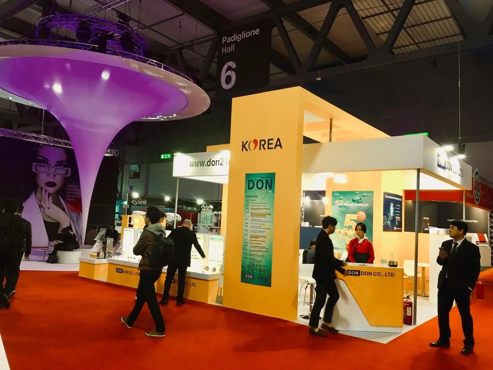Stand fieristico con il nome 'KOREA' e 'DON CO., LTD.' mostrato sopra, persone che parlano e camminano, ambientato in una fiera con pavimentazione rossa.