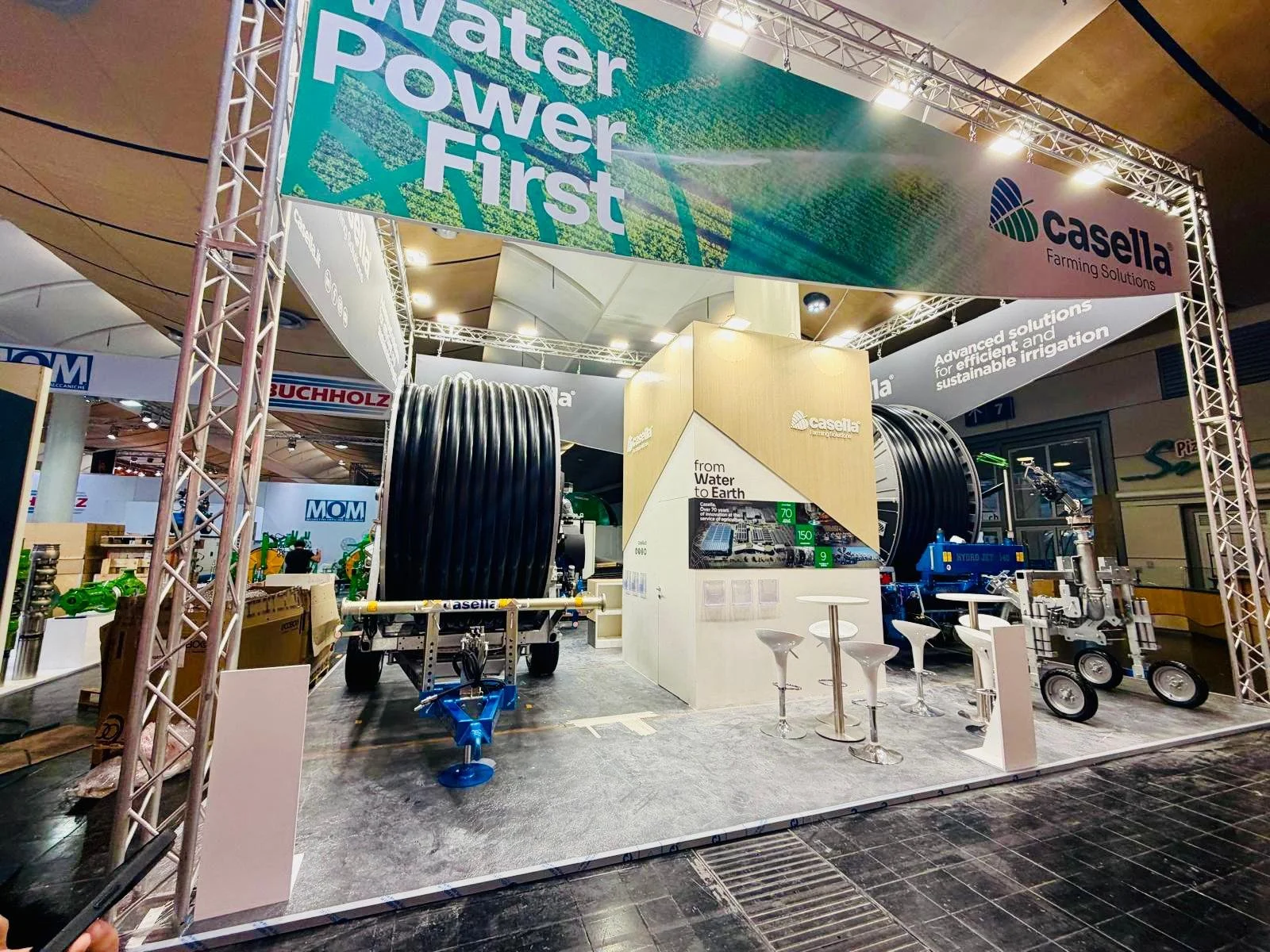 Stand espositivo di Casella Farming Solutions con attrezzature agricole come tubi neri di grandi dimensioni, macchinari e sedie bianche in un centro fieristico. Il cartello sopra recita 'Water Power First'.