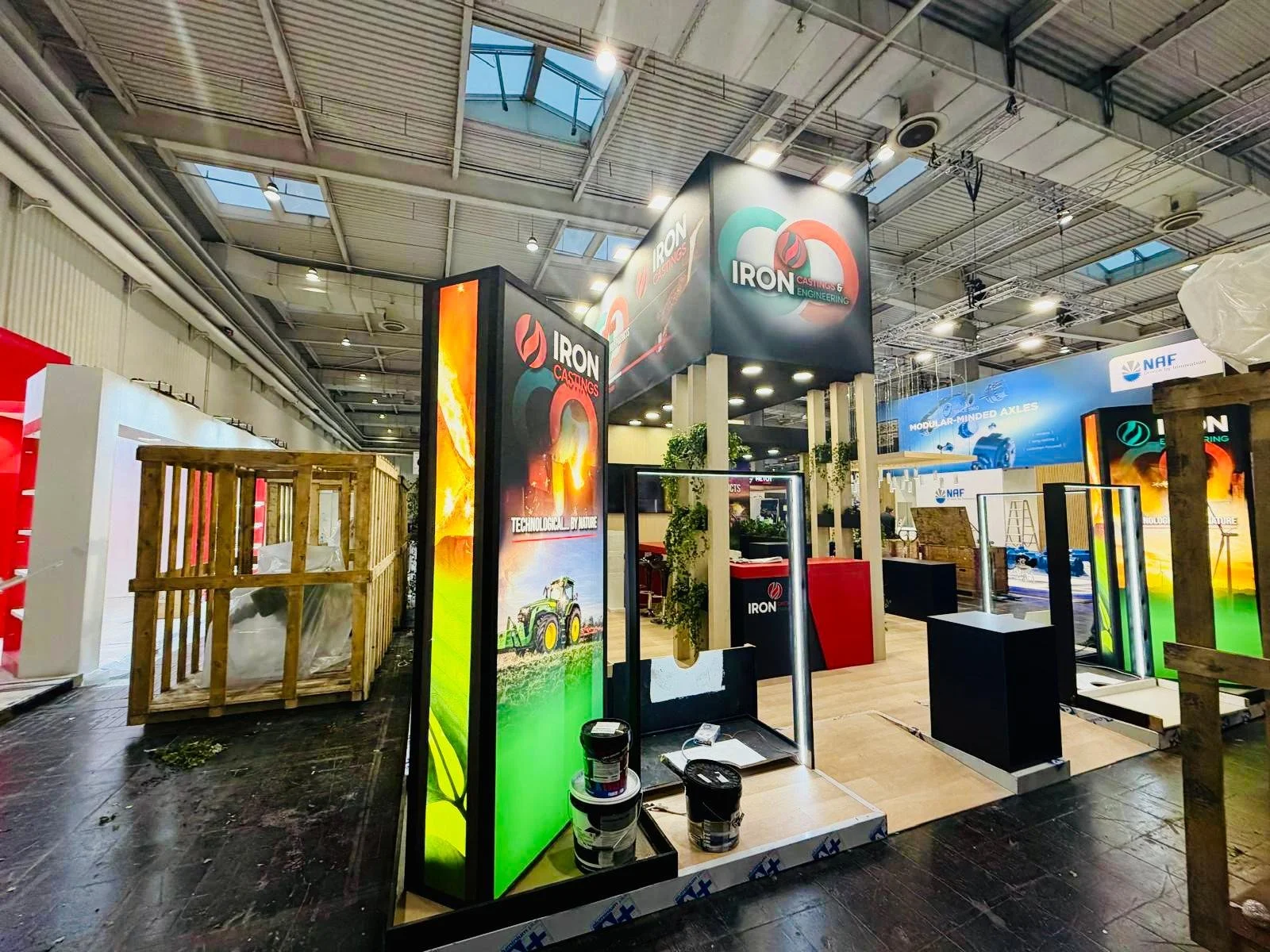 Stand fieristico con segnaletica e pannelli pubblicitari di Iron Castings, ambiente interno con soffitti alti e luci, decorazioni con piante, e strutture di legno.