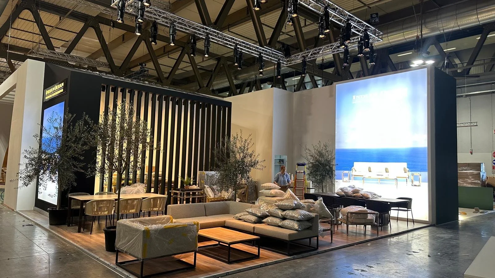 Stand fieristico con arredamento da esterno, divani, tavoli e piante, con grande schermo che mostra il mare sul retro.