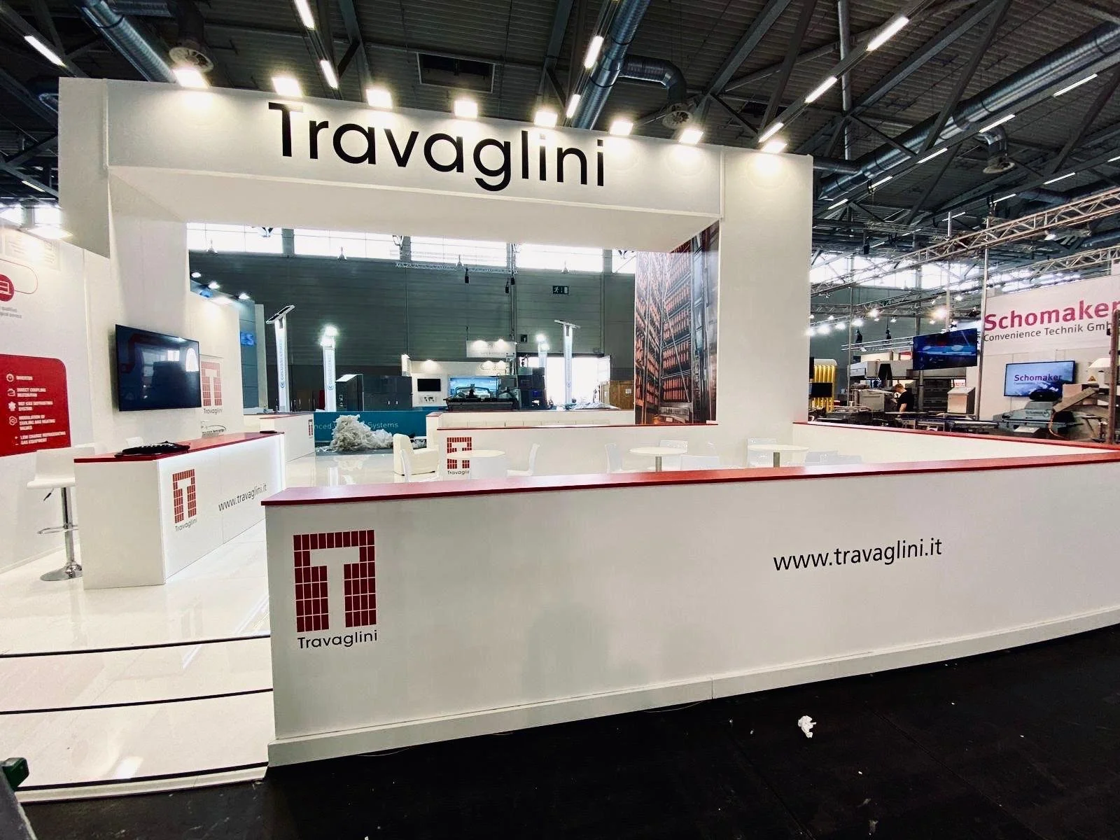 Stand espositivo con il nome 'Travaglini' in una fiera o esposizione, con una barriera bianca e logo aziendale, arredi bianchi e sfondo strutture del padiglione fieristico.