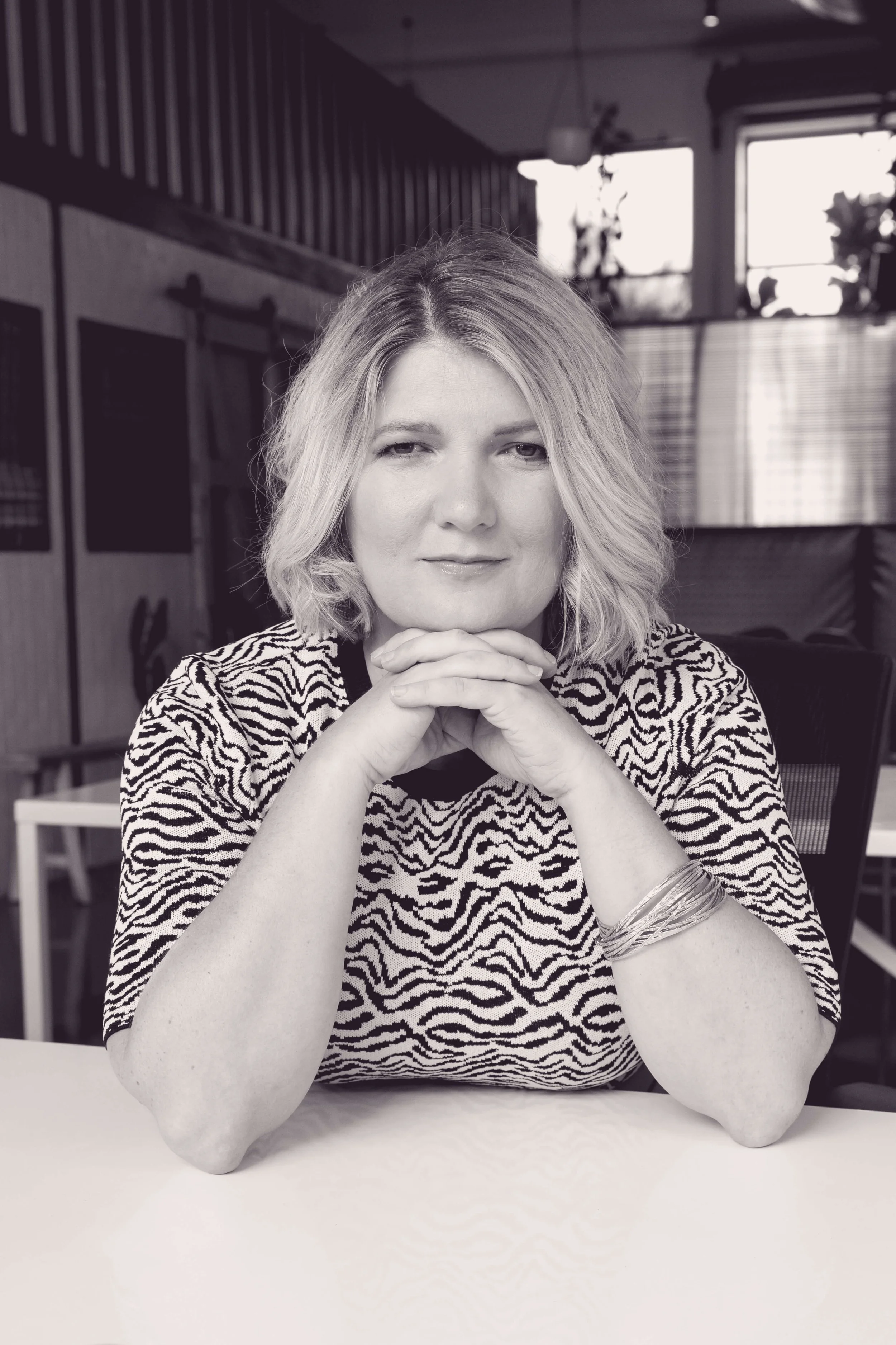 A woman with shoulder-length blonde hair sitting at a table, resting her chin on her clasped hands, wearing a zebra print top, in a cafe with drinks and plants in the background.