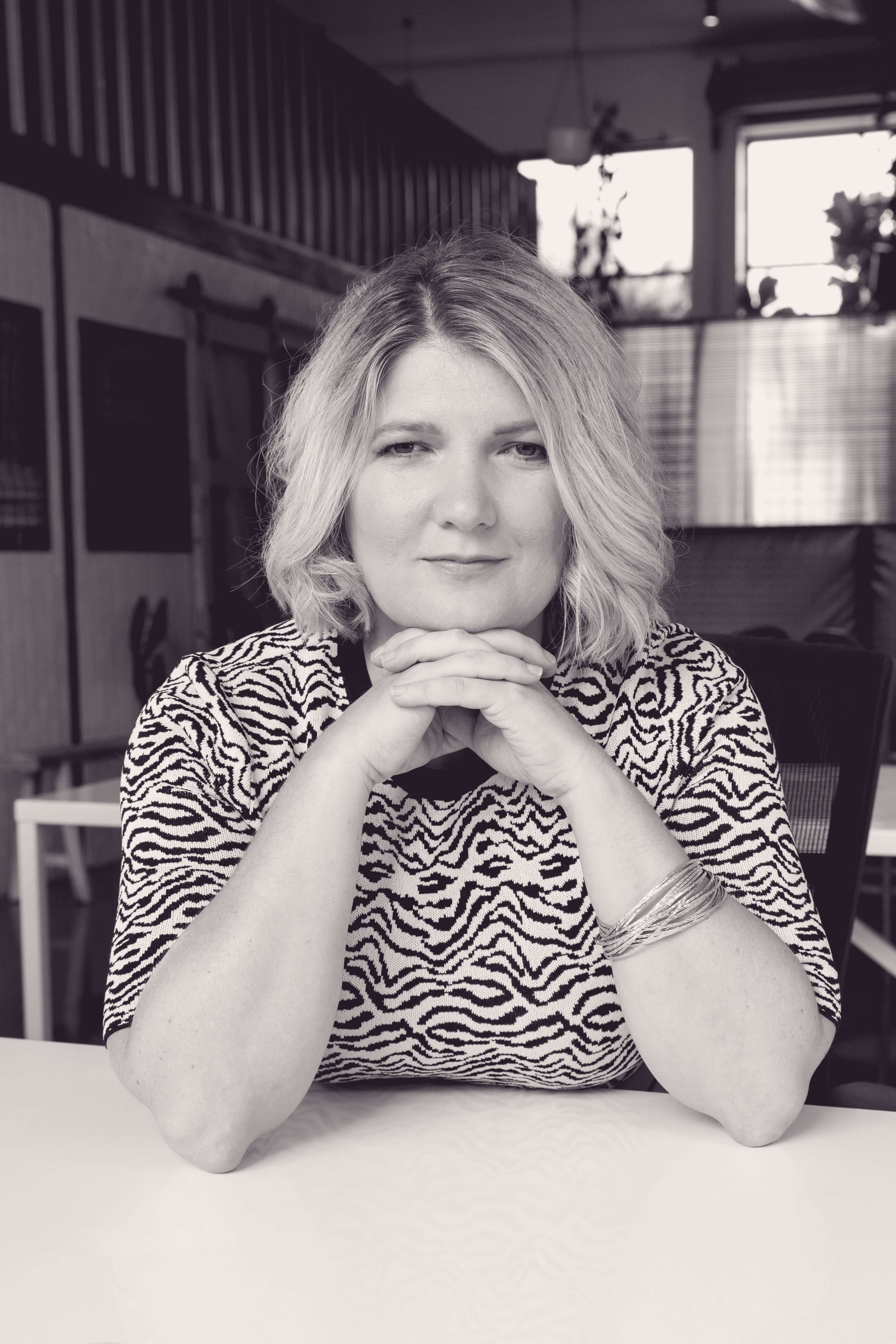 Black and white photo of a woman sitting at a table with her hands clasped under her chin, wearing a patterned top and a bracelet, in a cozy indoor setting with windows and plants in the background.
