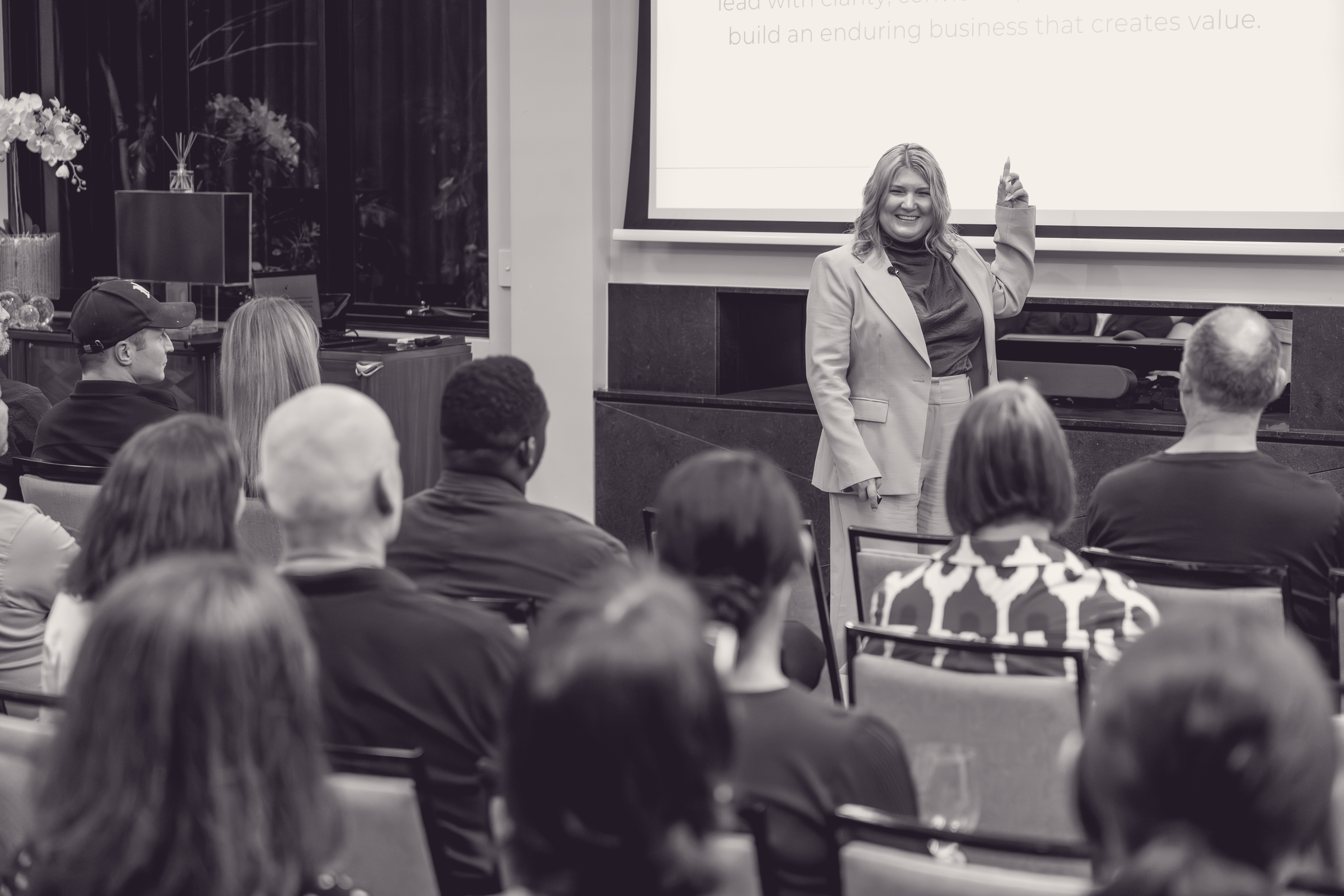 A woman in a blazer giving a presentation to a seated group in a conference room.