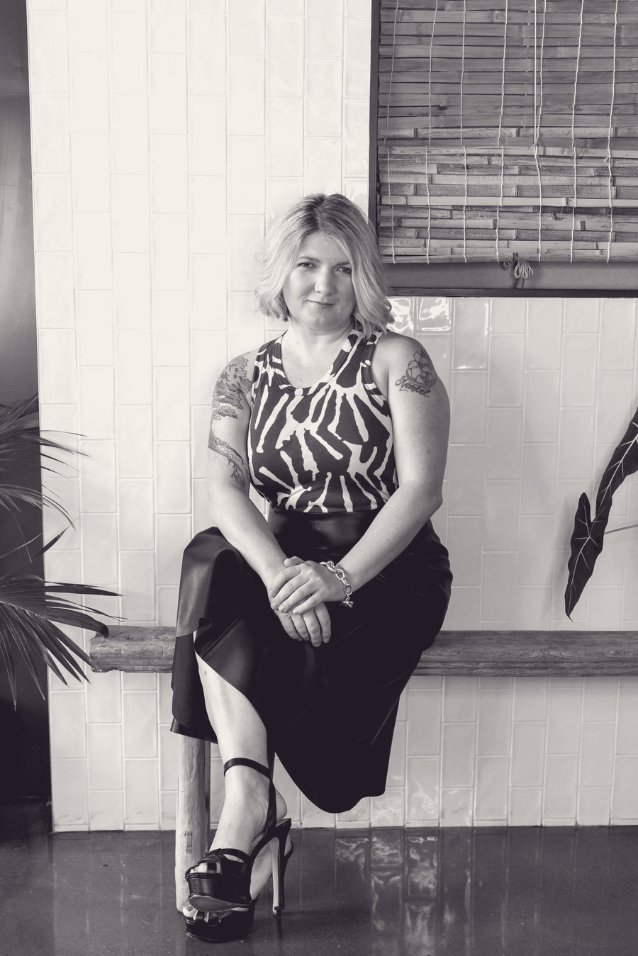 A woman with shoulder-length blonde hair sitting on a wooden bench indoors. She is wearing a sleeveless animal print top, a black skirt, and high heels. She has tattoos on her arms and wears jewelry. The background includes a tiled wall, a window with blinds, and some plants.