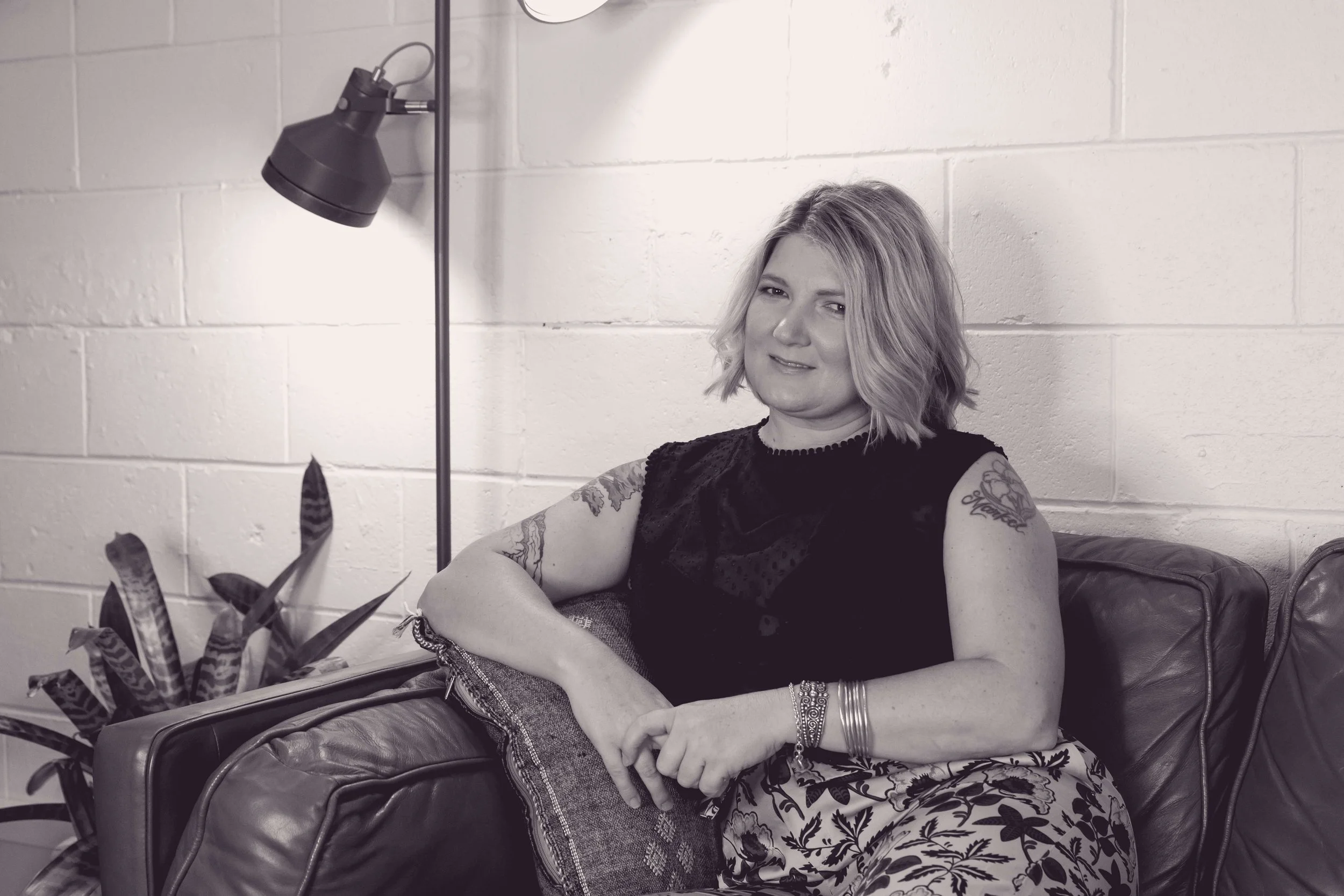A woman with shoulder-length wavy hair, tattoos on her arms, and wearing a black top, sitting on a couch in a room with white brick walls. There's a lamp and a plant in front of her.