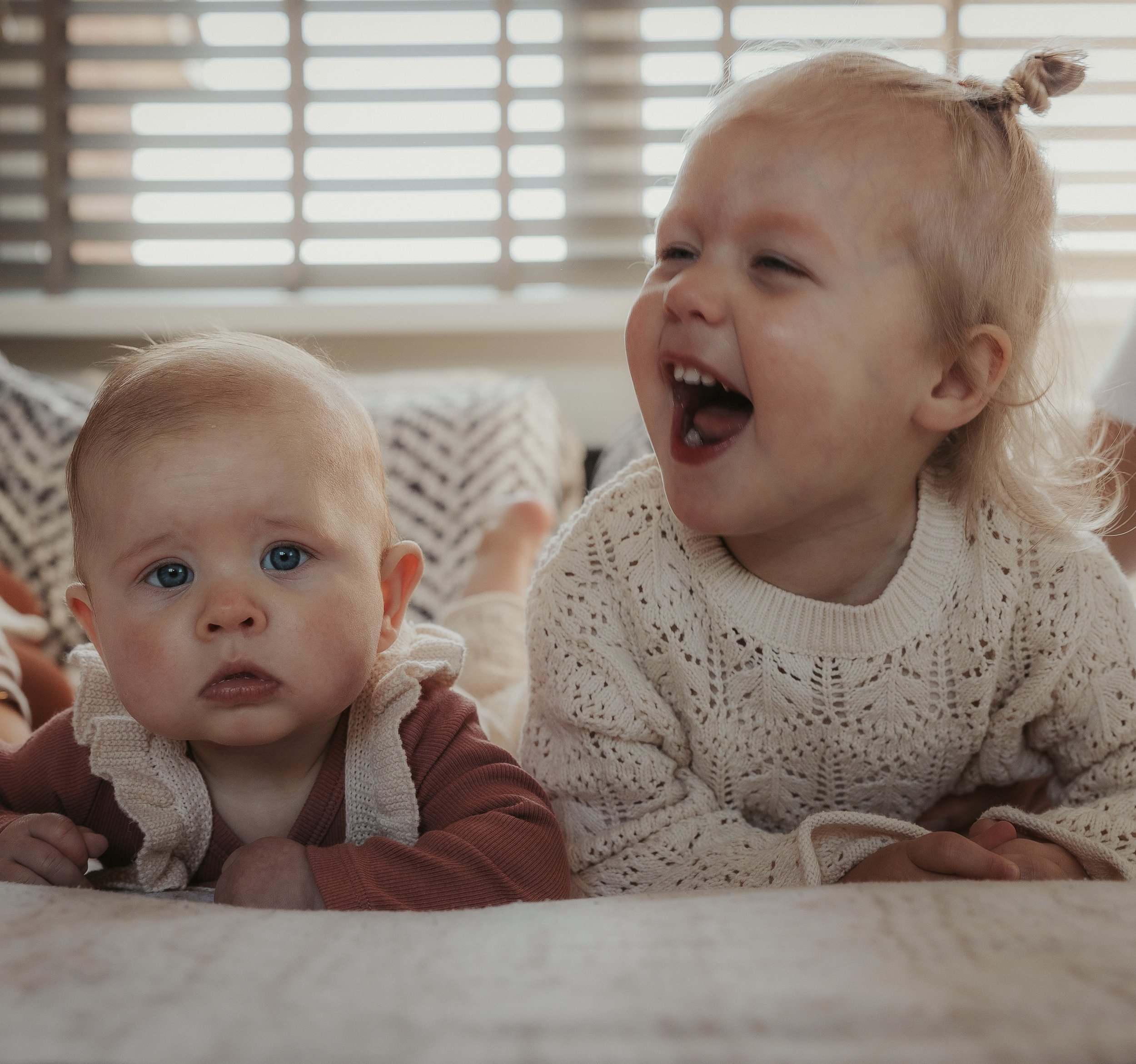 Twee jonge kinderen liggen op de vloer binnen, één kijkt serieus naar de camera en de andere lacht enthousiast.