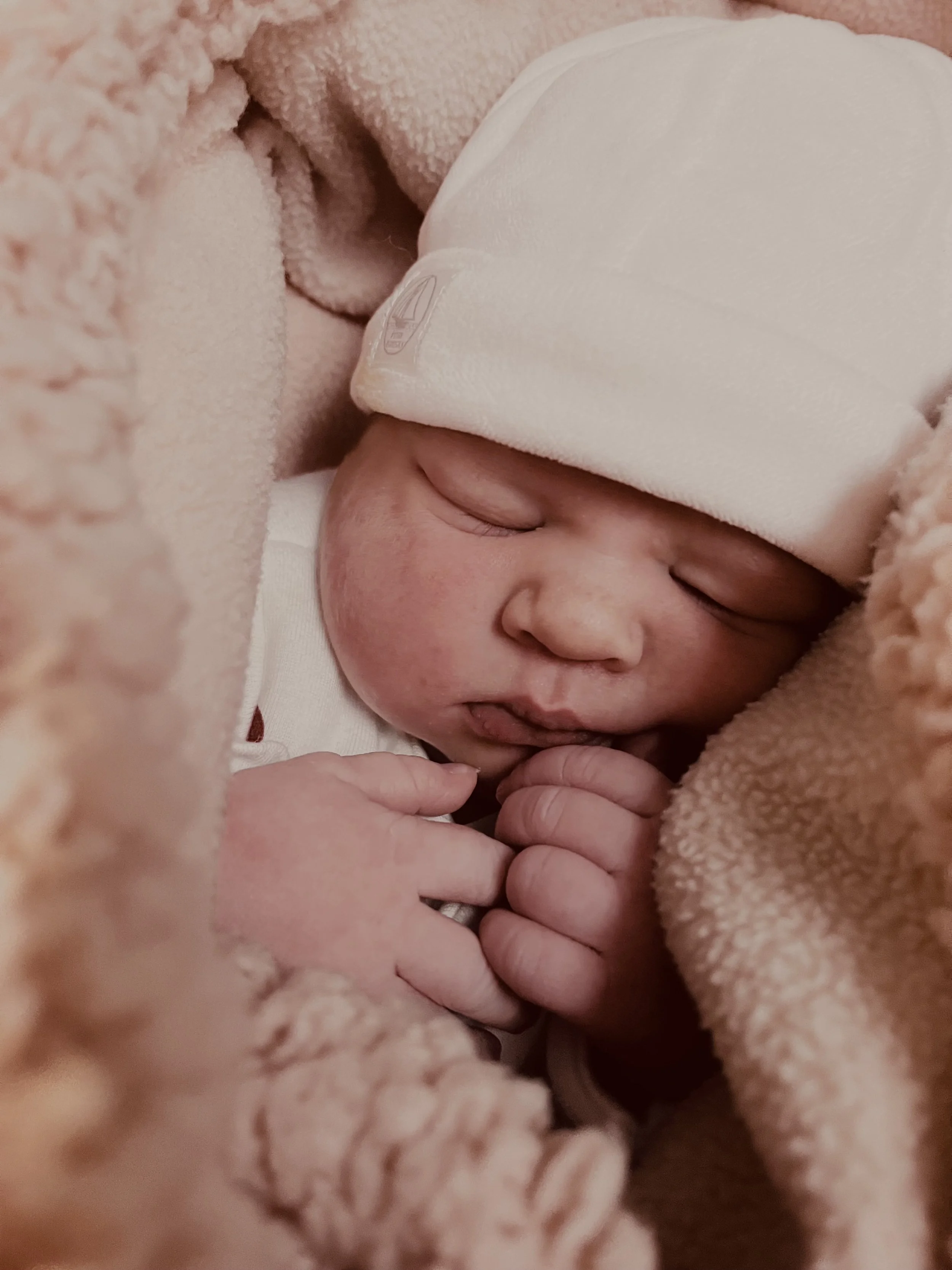 Een pasgeboren baby ligt ingepakt in een zachte, roze deken en draagt een witte muts, slapend met handjes dicht tegen het gezicht.