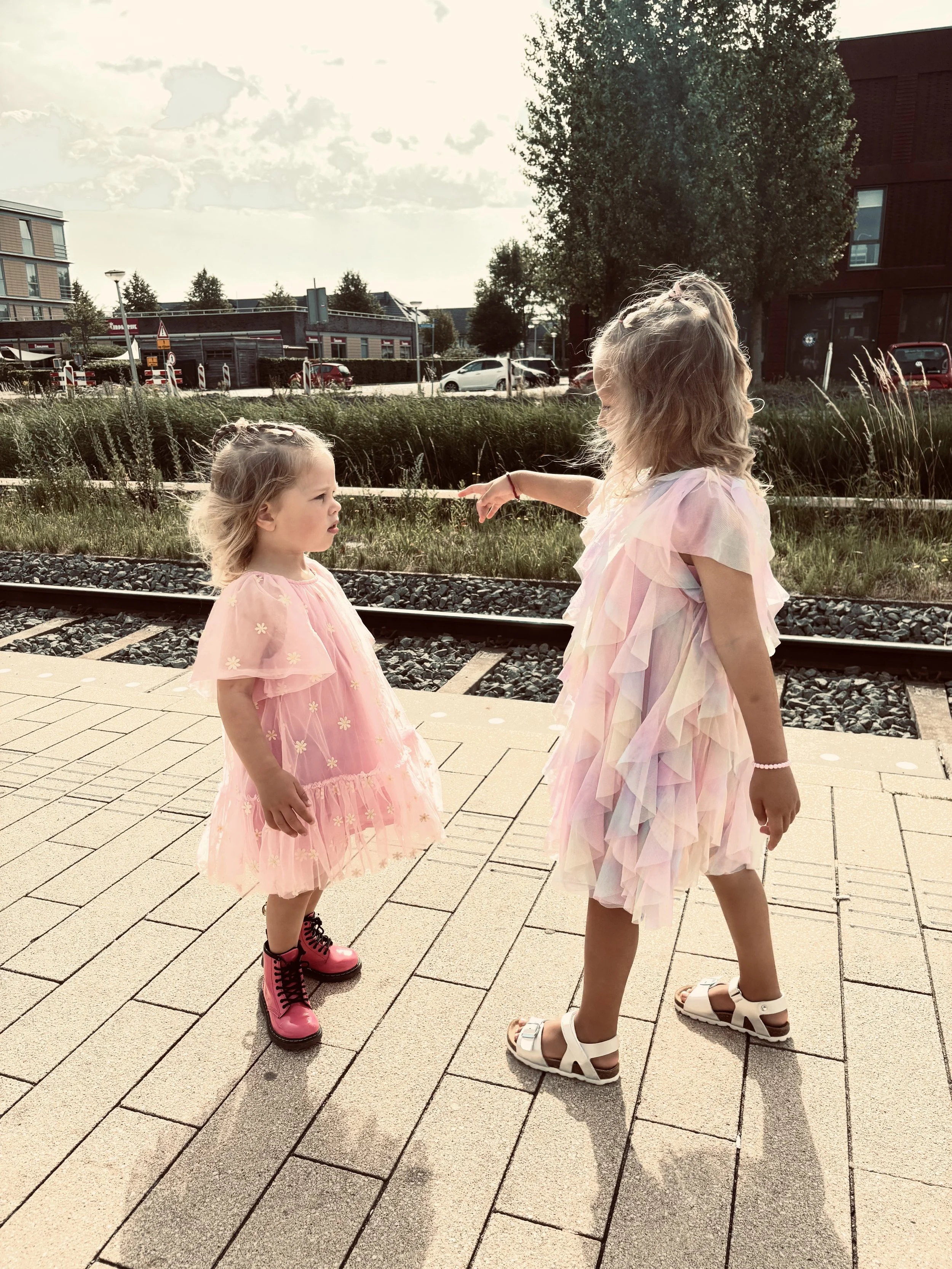 Twee jonge meisjes in pastelgekleurde jurkjes staan op een perron naast een spoorlijn, de ene meisjes wijst naar de ander.