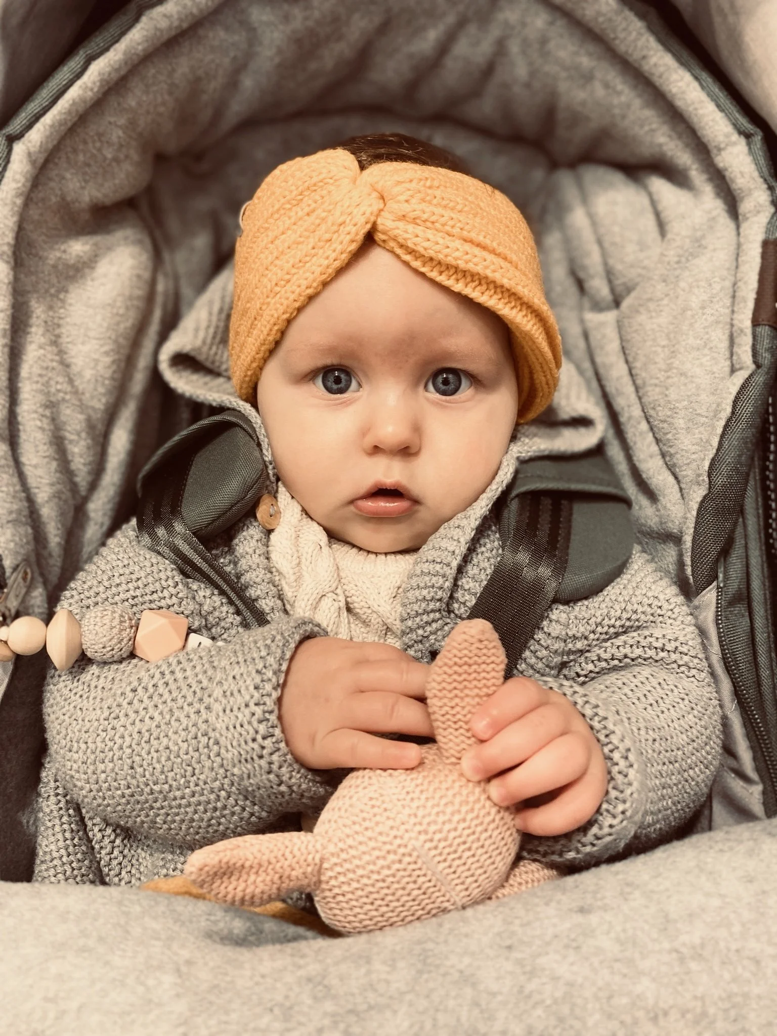 Een baby met blauwe ogen, gekleed in een grijs gekleurd trui en een oranje gebreide hoofdband, die een knuffel vasthoudt, zittend in een kijkwieg.
