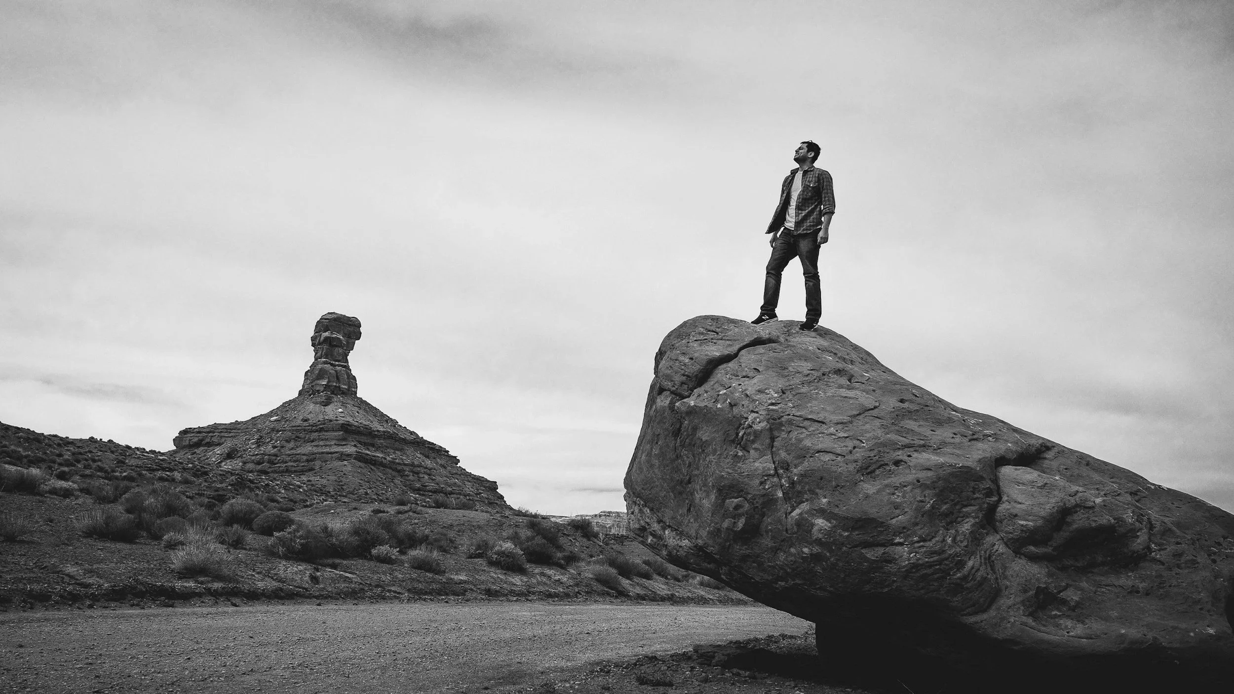 A man stands atop a large rock in a desert landscape, with a rock formation in the background. The image is in black and white.