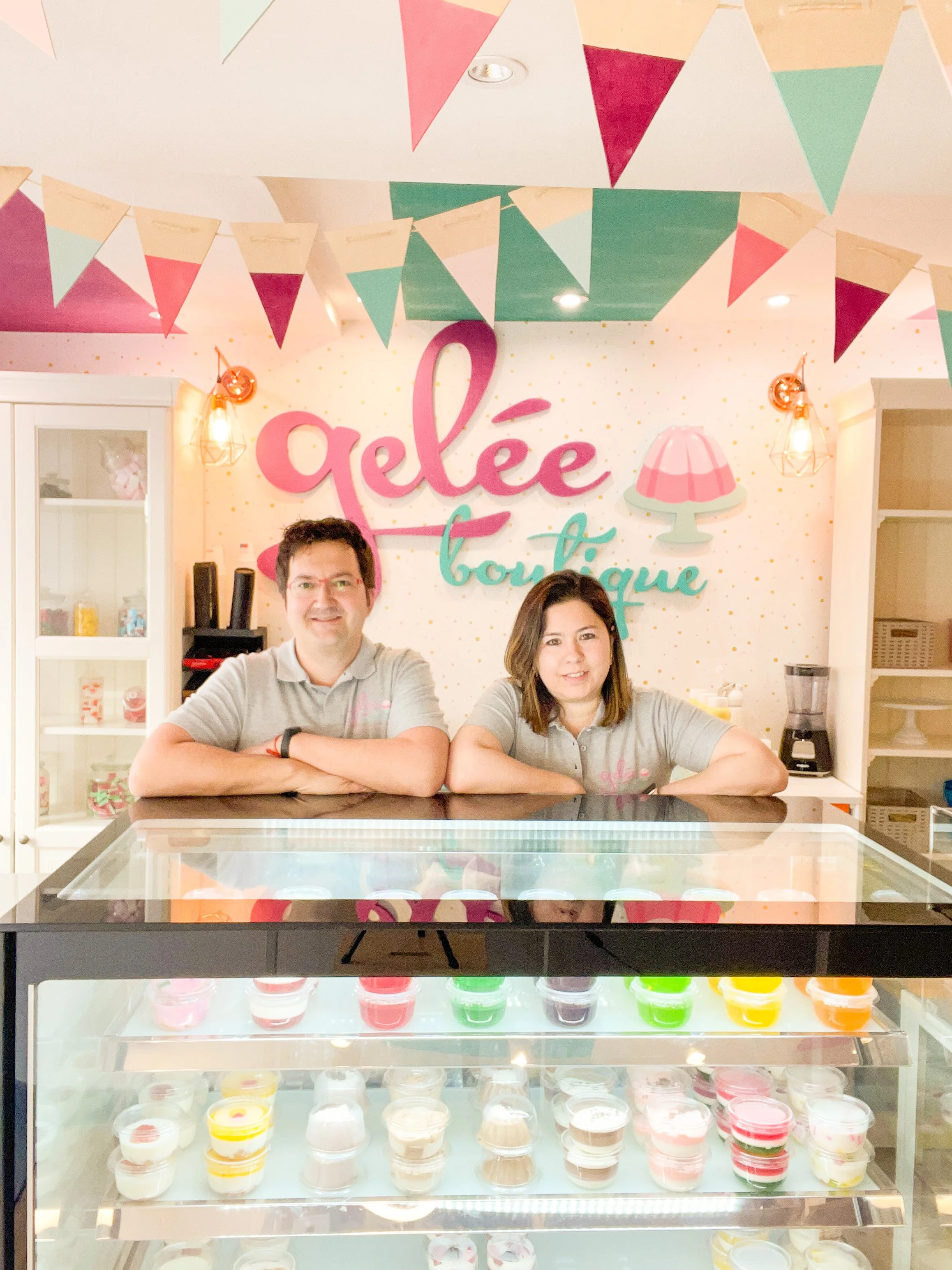 Dos personas detrás de un mostrador en una tienda de gelatinas y postres, decorada con banderines de colores y un cartel con el texto 'gelée boutique' en el fondo.