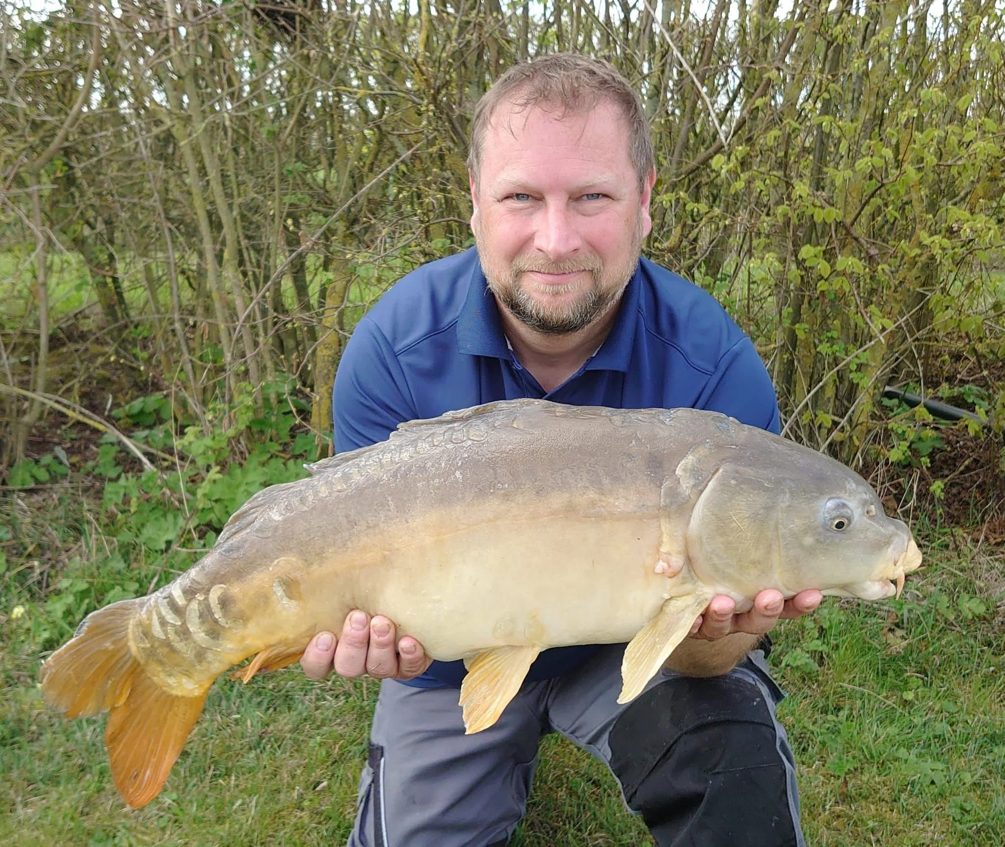 Man with short hair and beard holding large fish outdoors in a wooded area.