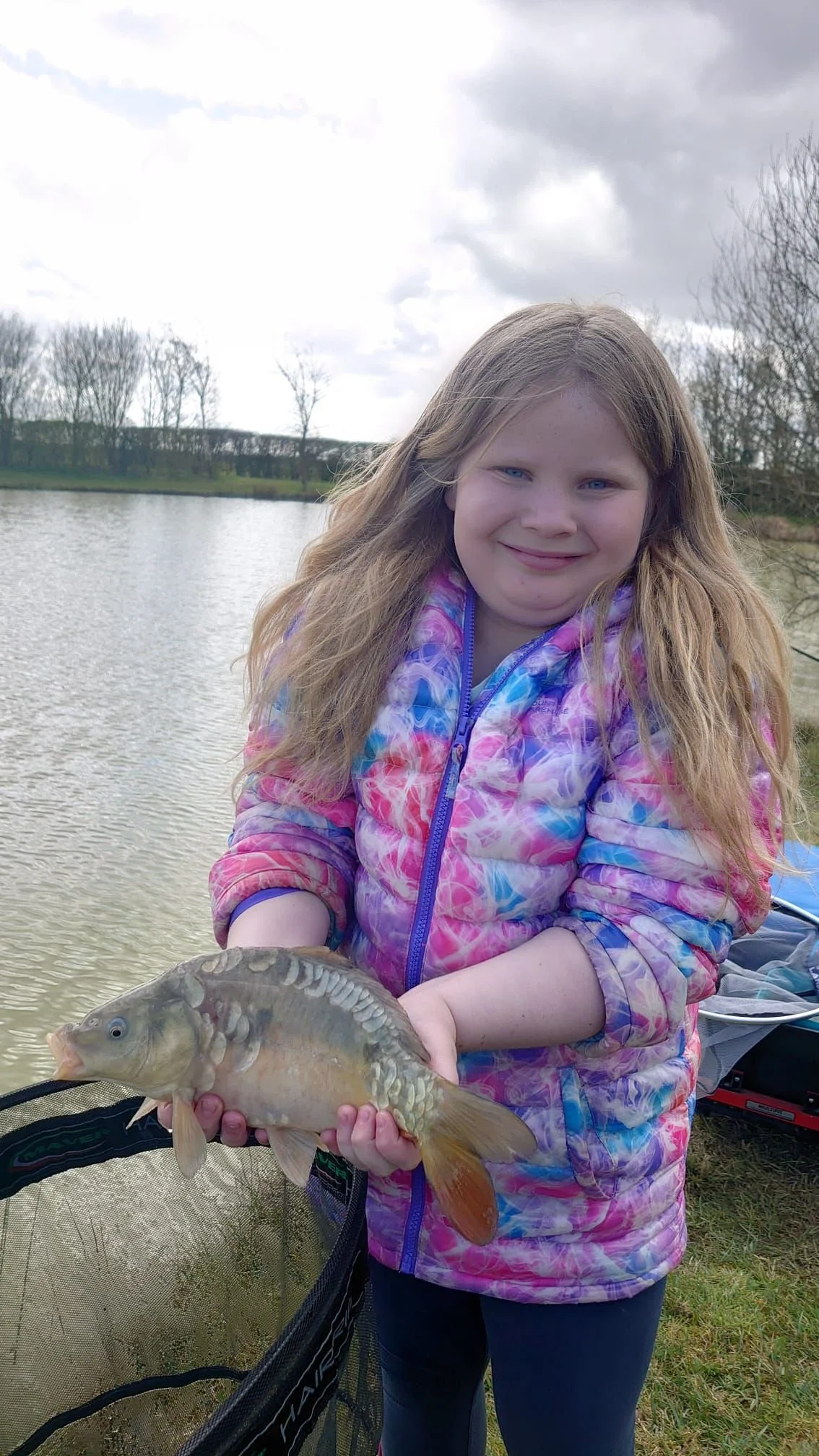 A girl holding a fish by a lake, smiling, wearing a colorful jacket, with overcast skies and trees in the background.