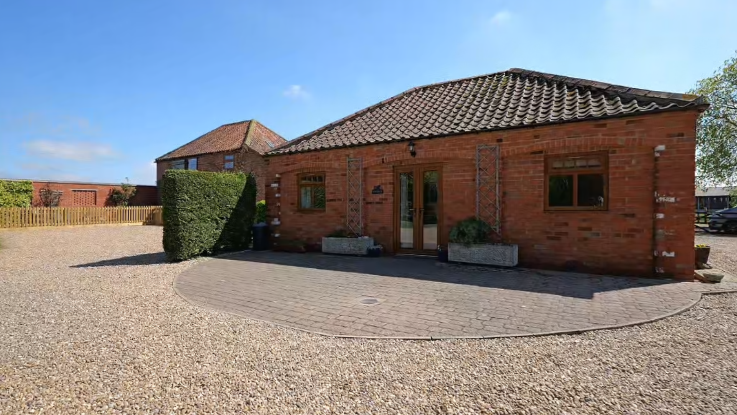 A brick building with a tiled roof, glass doors, and windows, surrounded by a gravel area and a small paved patio, with a blue sky above.