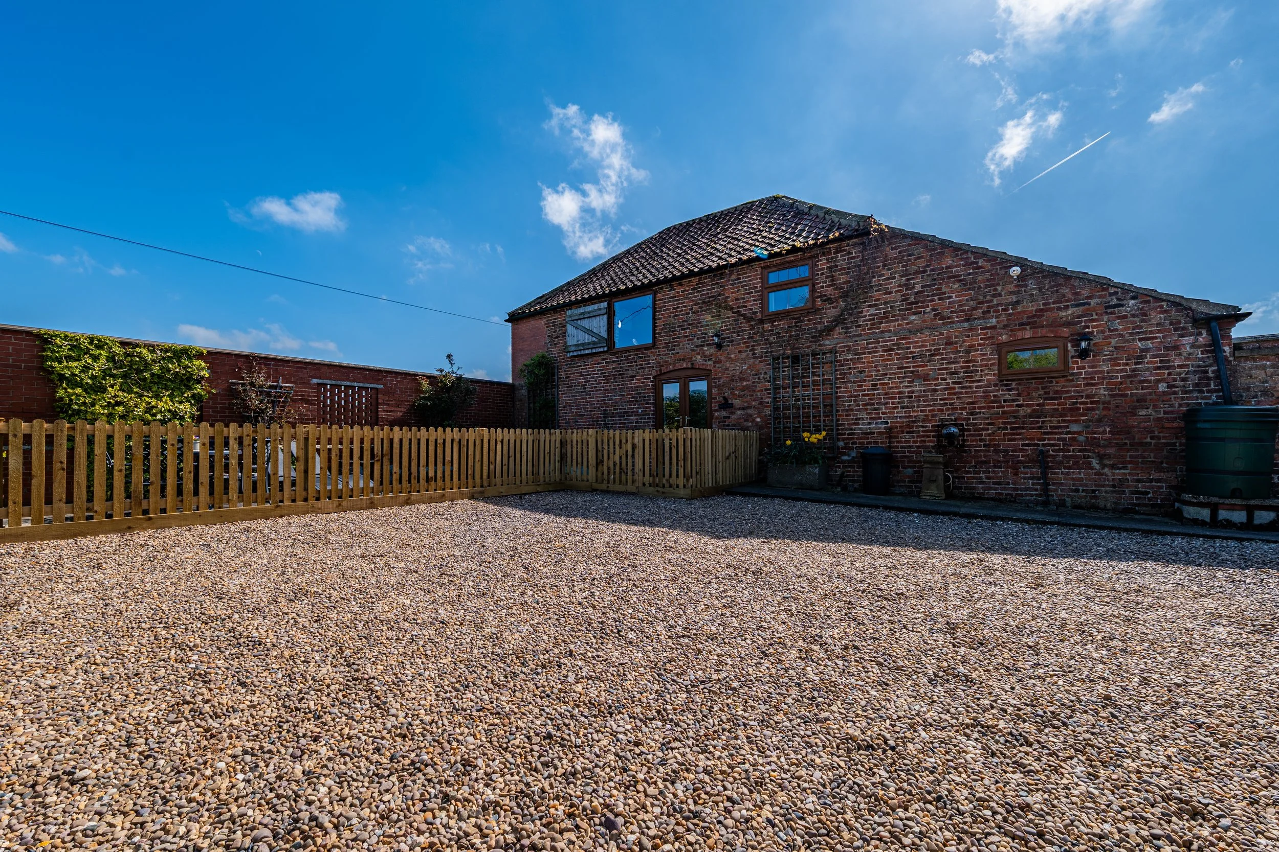 A brick house with a tiled roof and small windows, surrounded by a wooden fence, under a blue sky with scattered clouds.