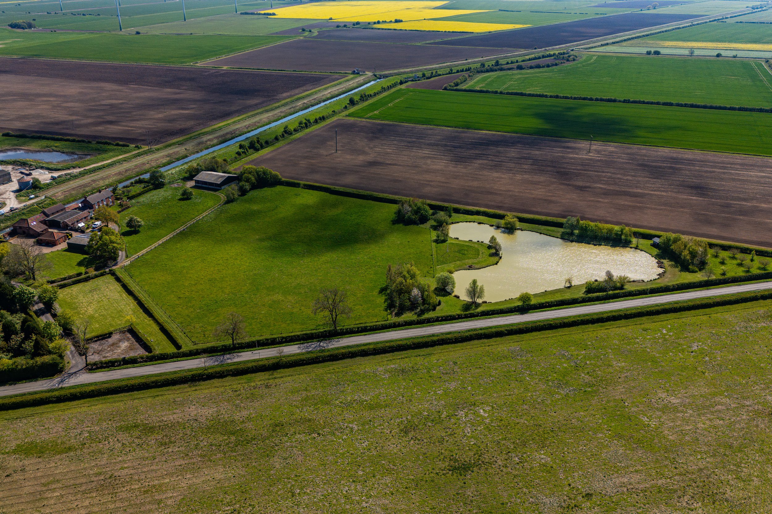 Aerial view of farmland with colorful fields, a canal lined with trees, residential houses, and a small pond, under bright daylight.