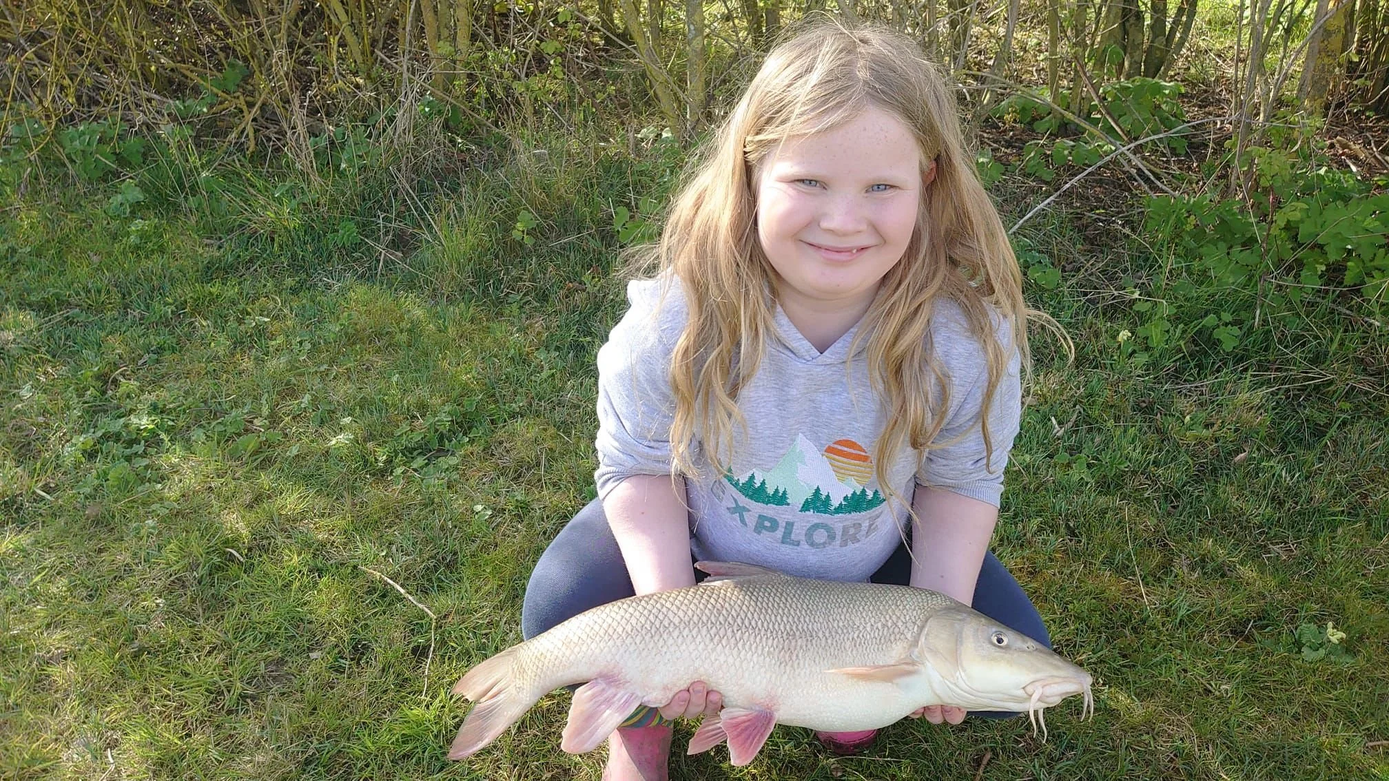 A young girl with long blonde hair, wearing a gray hoodie with a graphic of mountains, trees, and a sun, is sitting on grass outdoors, holding a large fish she caught.