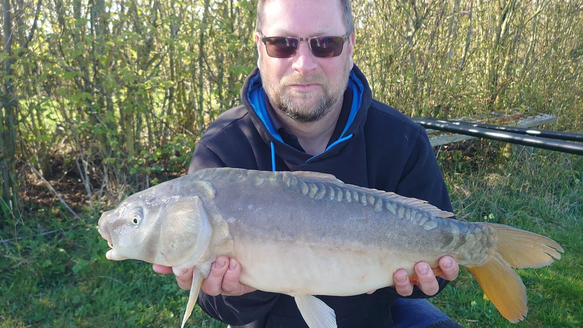 A man wearing sunglasses and a black jacket holds up a large fish outdoors with green trees in the background.
