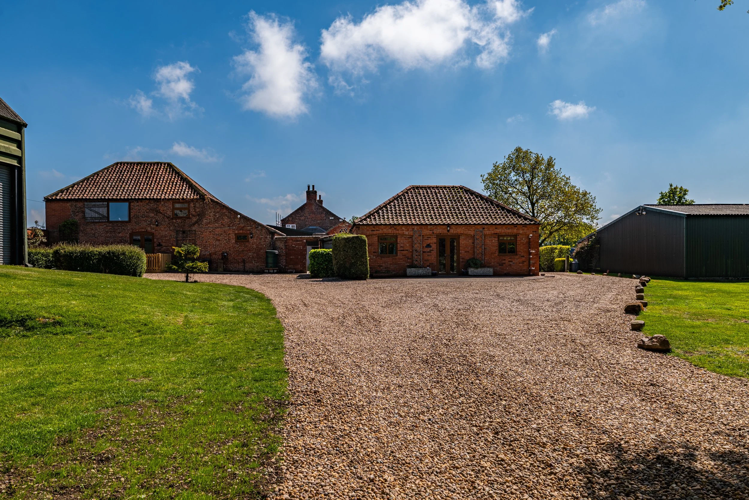 View of three brick houses with tiled roofs, a gravel driveway, green lawn, bushes, and trees under a partly cloudy blue sky.