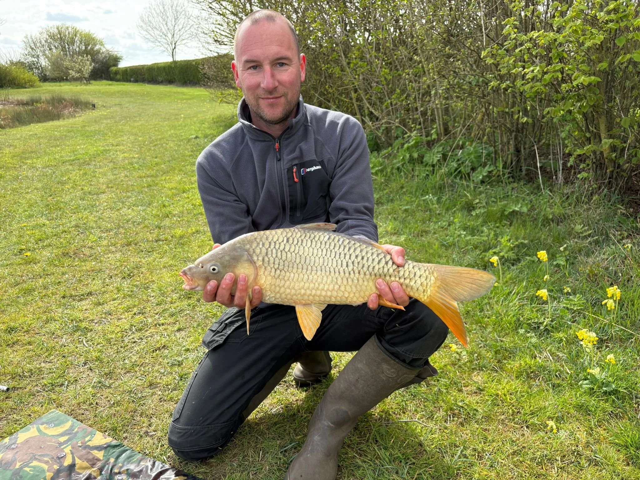 A man kneeling on grass outdoors holding a large fish, with trees and bushes in the background on a cloudy day.