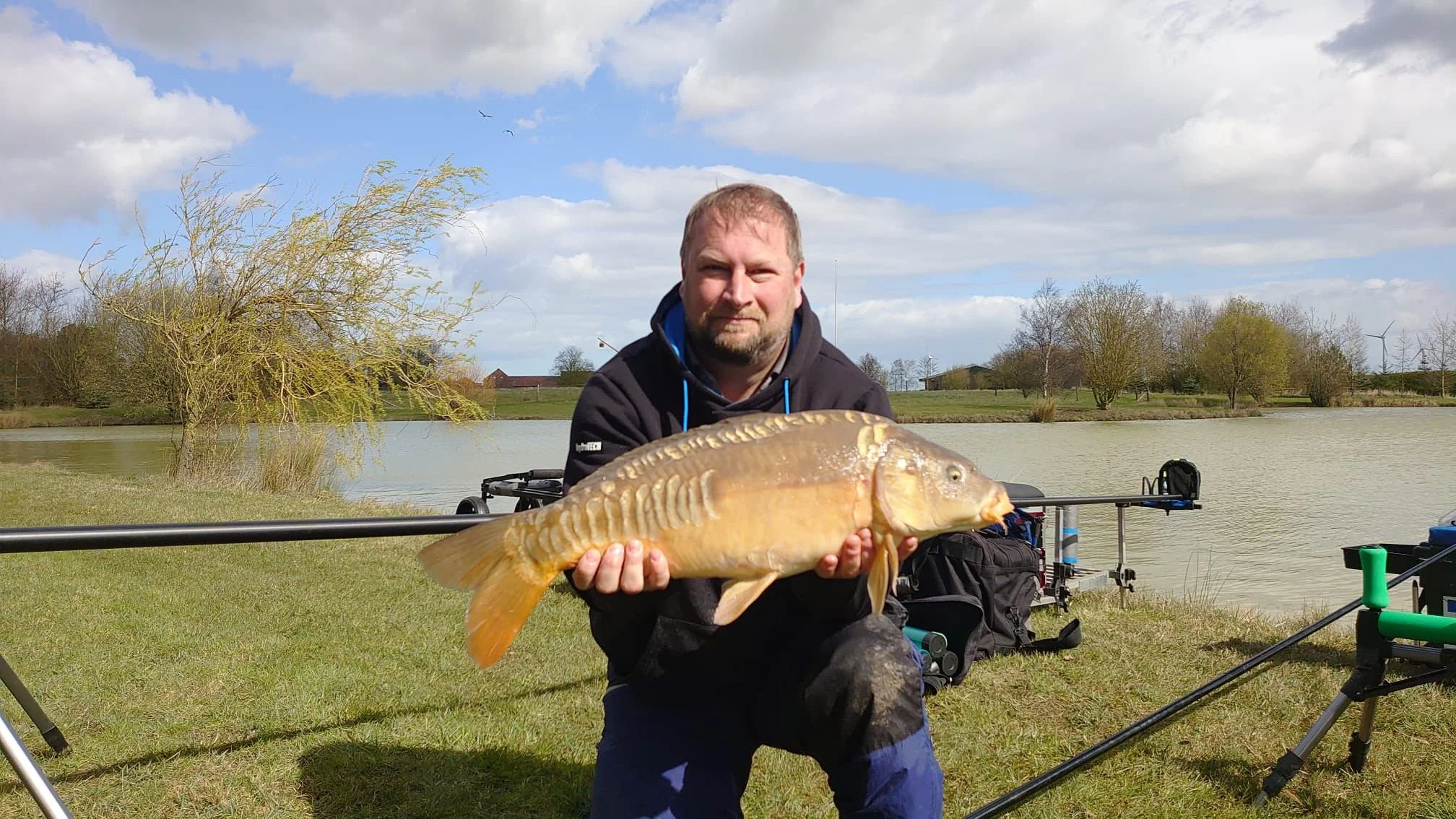 A man kneeling by a lake holding a large fish with both hands, there are fishing gear and a wind turbine in the background, under a partly cloudy sky.