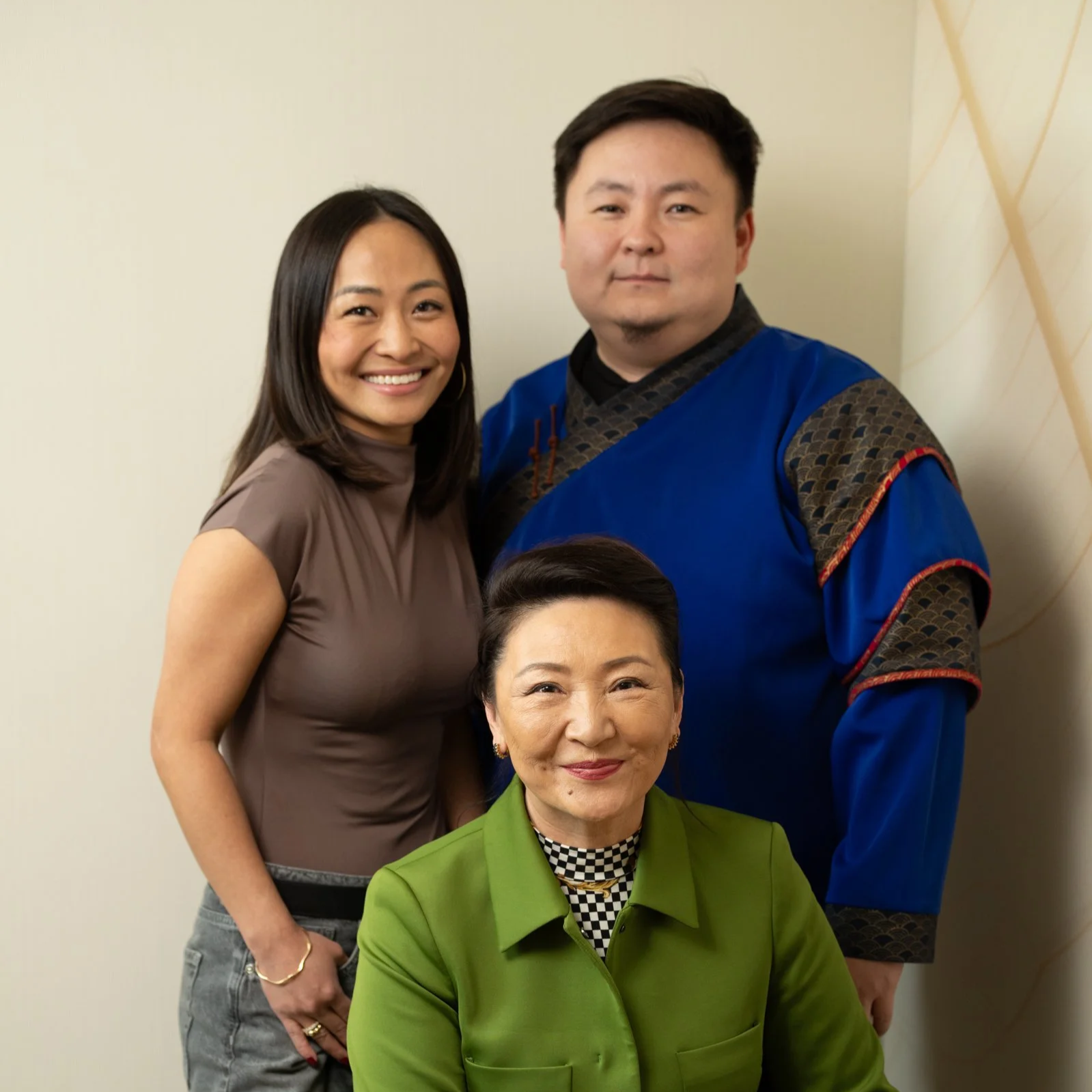 Three people posing for a photo, smiling, in front of a light-colored wall. Two women are standing, one seated. The woman in the front is wearing a green jacket, the woman standing is in a taupe top, and the man standing next to her is wearing a traditional blue garment with black and red accents.