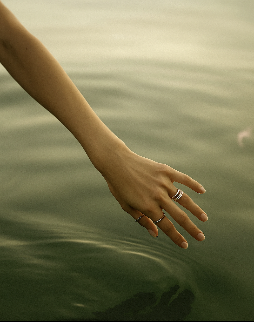 A person's arm and hand reaching into calm, greenish water, with multiple rings on fingers.