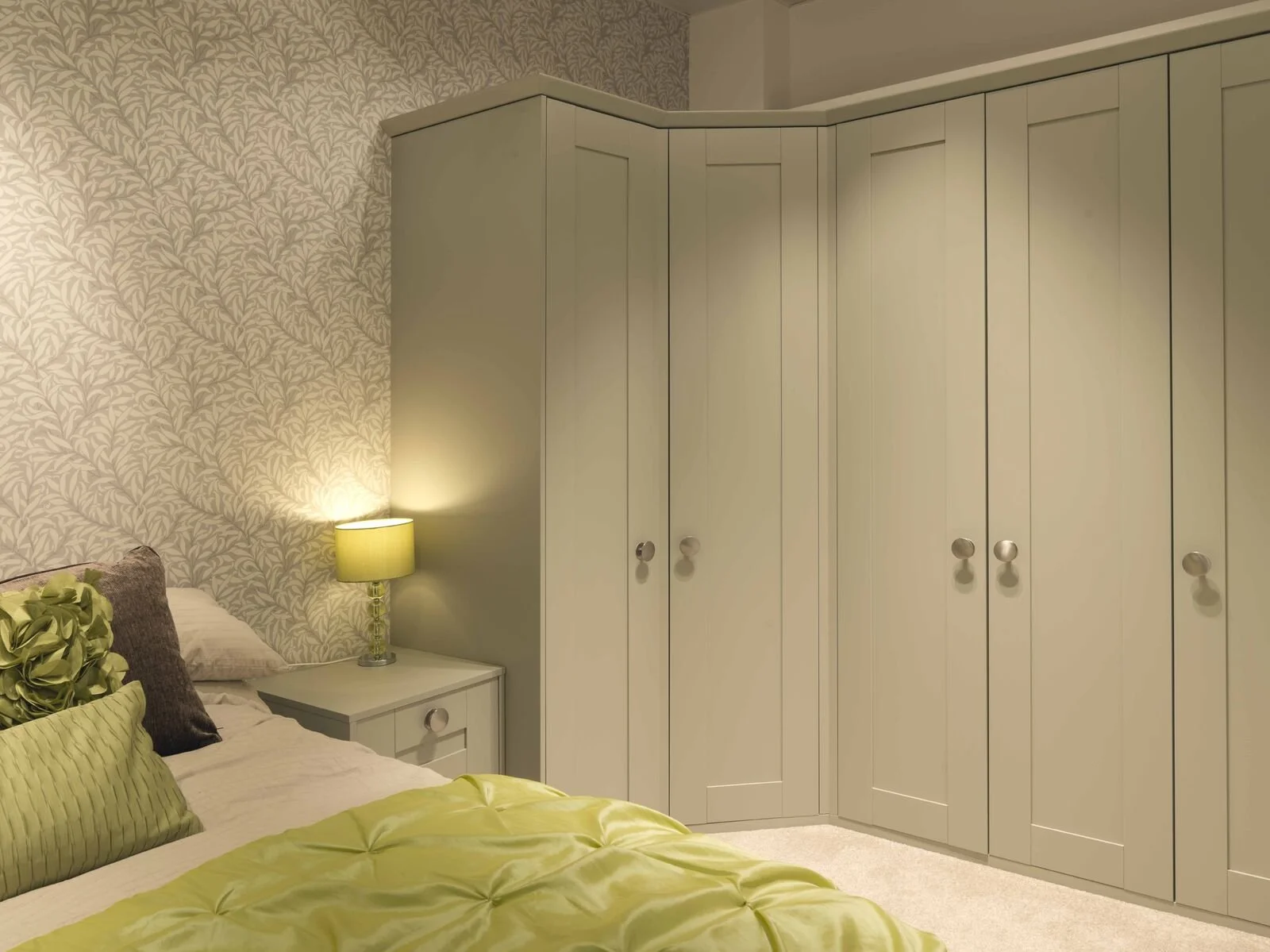 A bedroom corner featuring a bed with green and beige pillows, a nightstand with a green lamp, and a large beige wardrobe with round knobs. The wall has a leafy patterned wallpaper.
