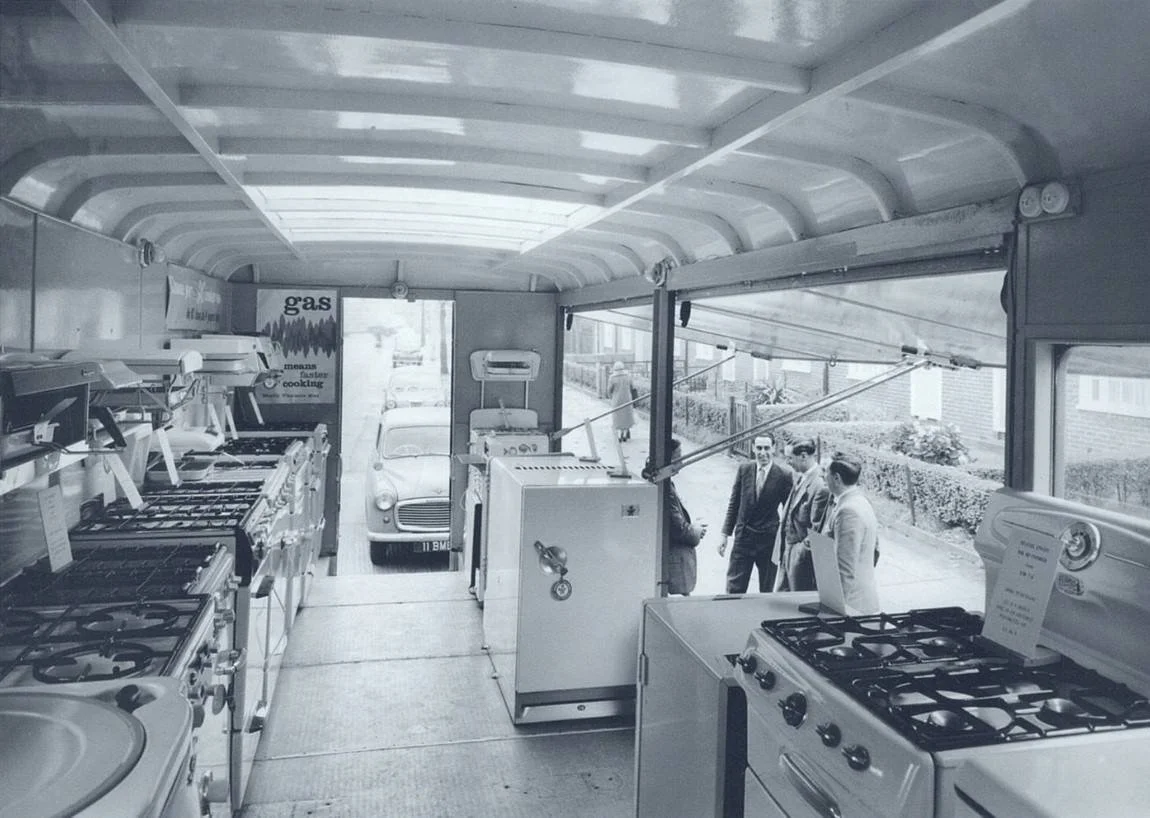 Black-and-white photo of a vintage store, likely an appliance shop, with stoves, ovens, and other appliances on display. Three men in suits are standing and conversing near the store entrance, while a woman walks past outside.