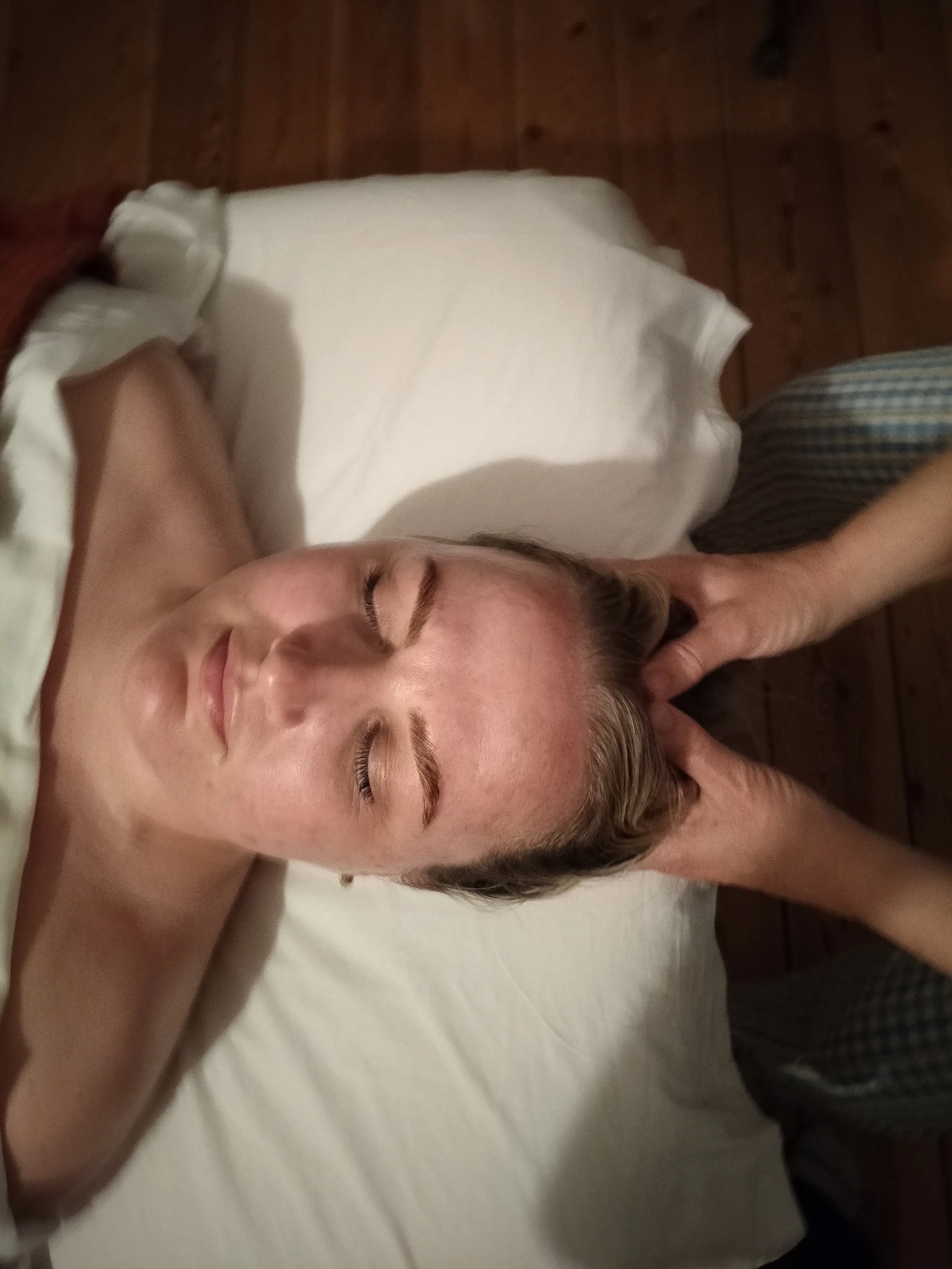 En kvinde ligger afslappet med lukkede øjne på en massagebriks, mens en person holder hænderne på hendes hoved.