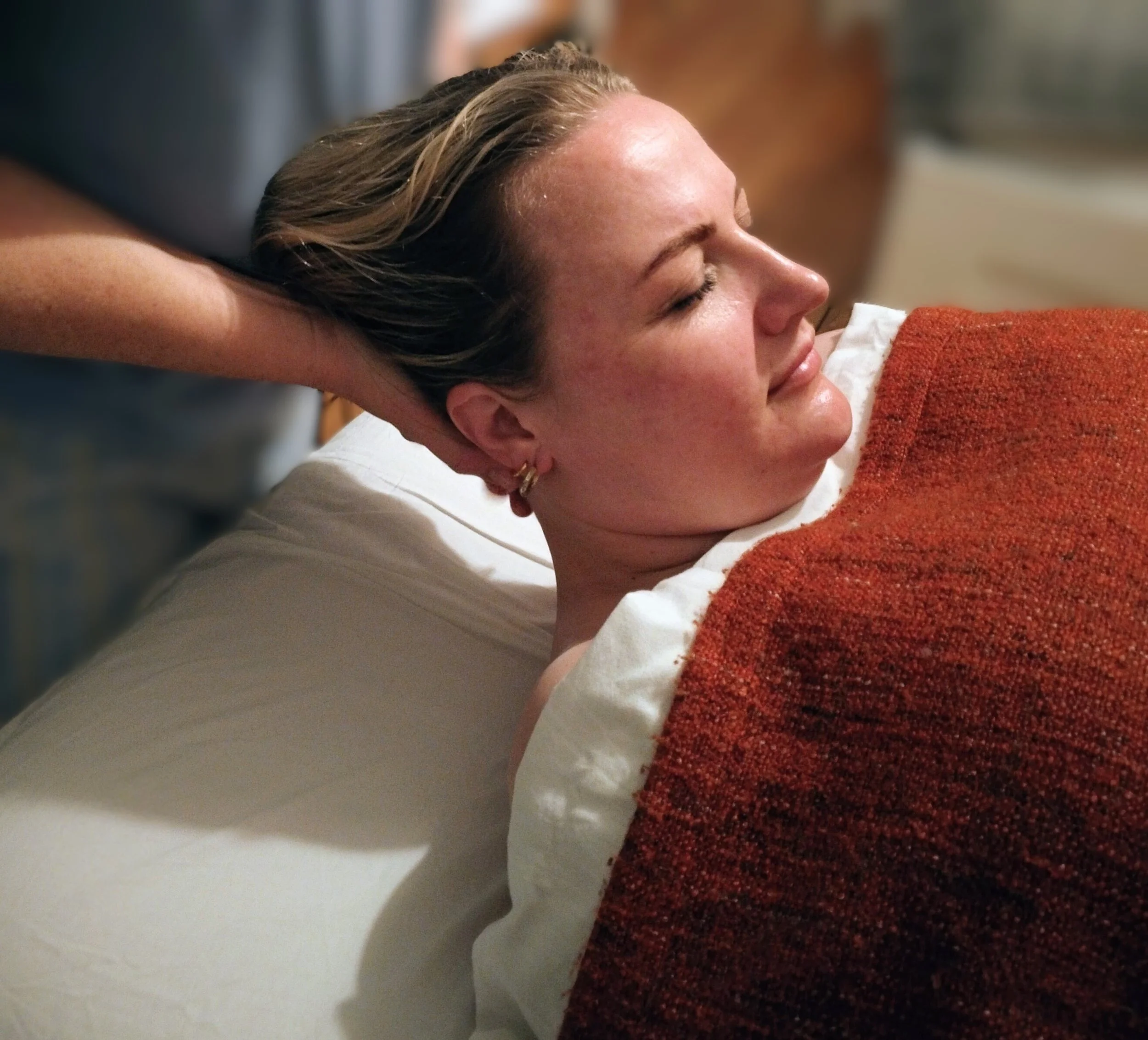 Kvindelig person ligger afslappet med lukkede øjne, modtager massage under kraniekanten, med et rødt tæppe over sig og et hvidt lagen under sig.
