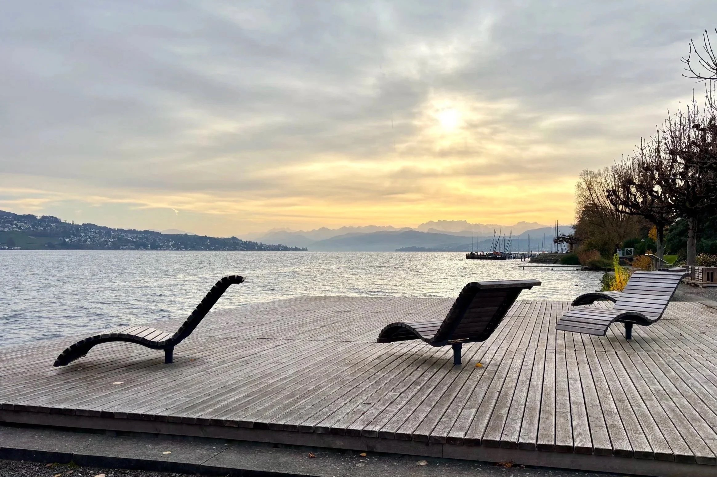 Sonnenaufgang über dem Zürichsee, mit Liegestühlen auf einer Holzterrasse im Vordergrund, Bäume und Boote am Ufer im Hintergrund, in Thalwil.